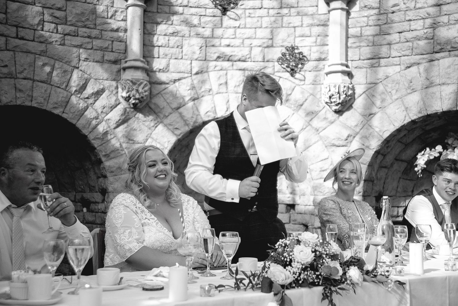Black and white photo of wedding guests enjoying dinner under stone arches.