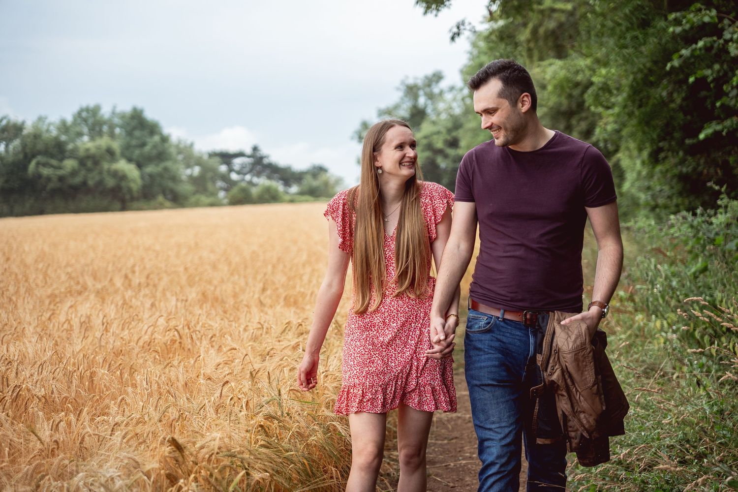 on the right couple walking holding hands and looking at each other along path next to a golden wheat field on the left