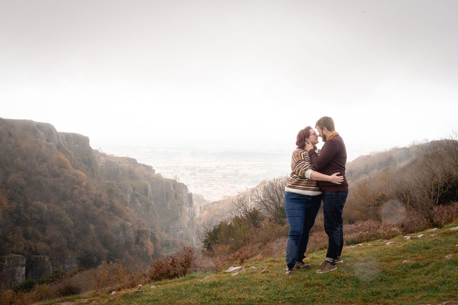 couple embracing on top of a hill on their engagement shoot, Cheddar gorge visible in the distance, mist and rain envelop the couple