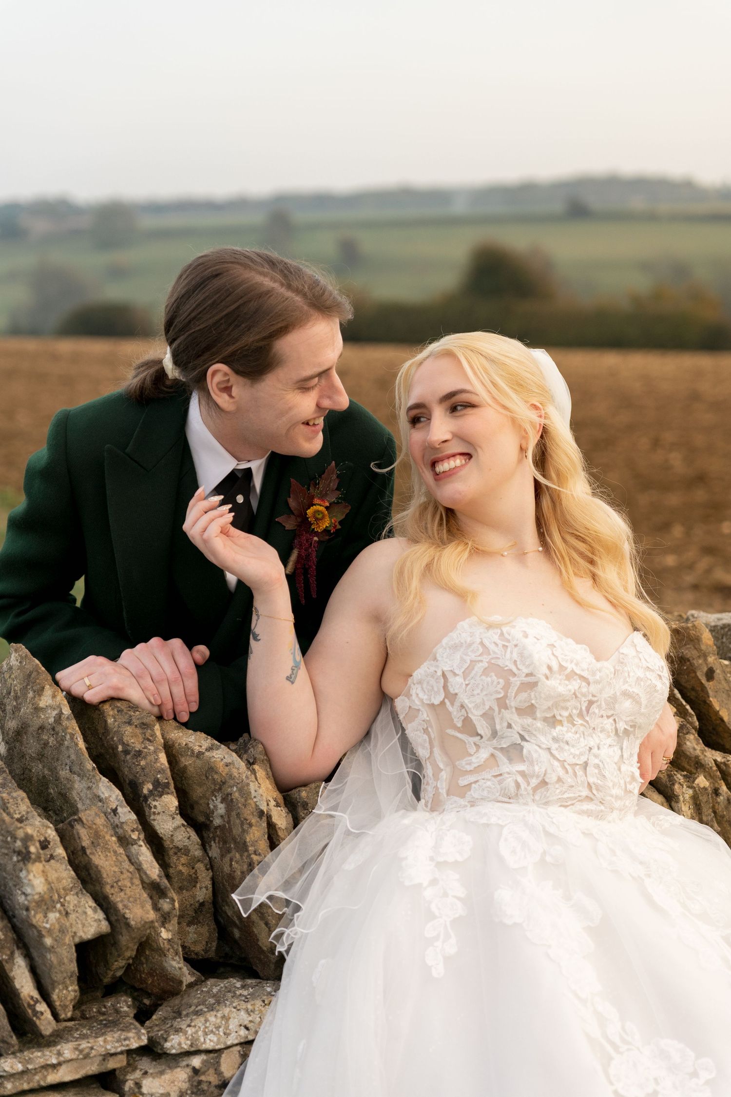 A smiling bride in a lace ballgown and groom in a dark green suit lean against a stone wall in a countryside setting.