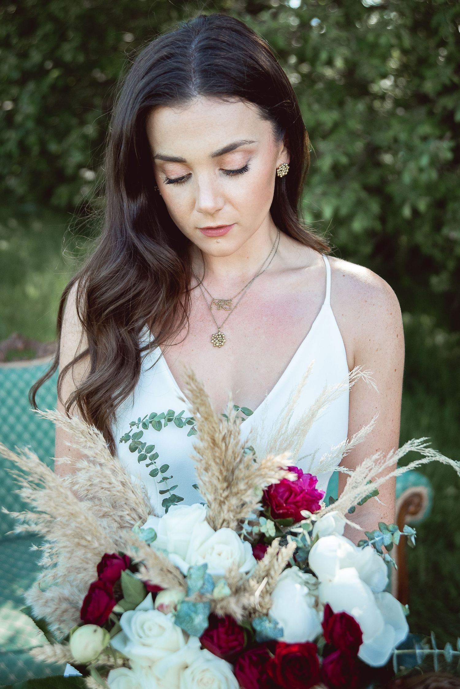 A bridal bouquet with white roses, deep red flowers, and dried pampas grass is held against a white dress.