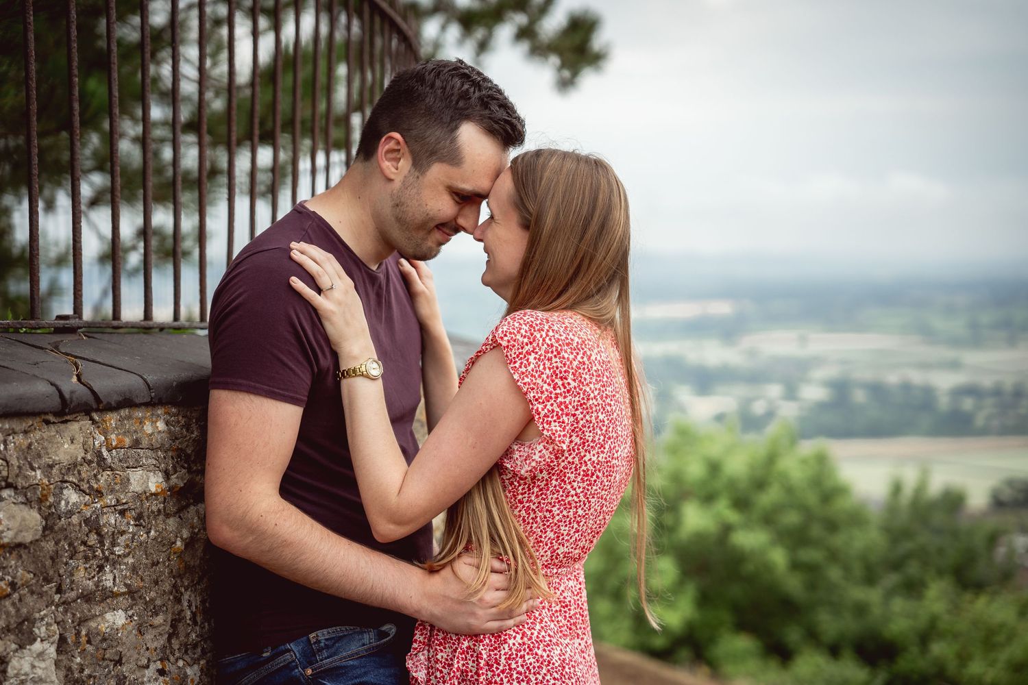 couple leaning against a wall and looking at each other, forehead to forehead, with scenery in the background