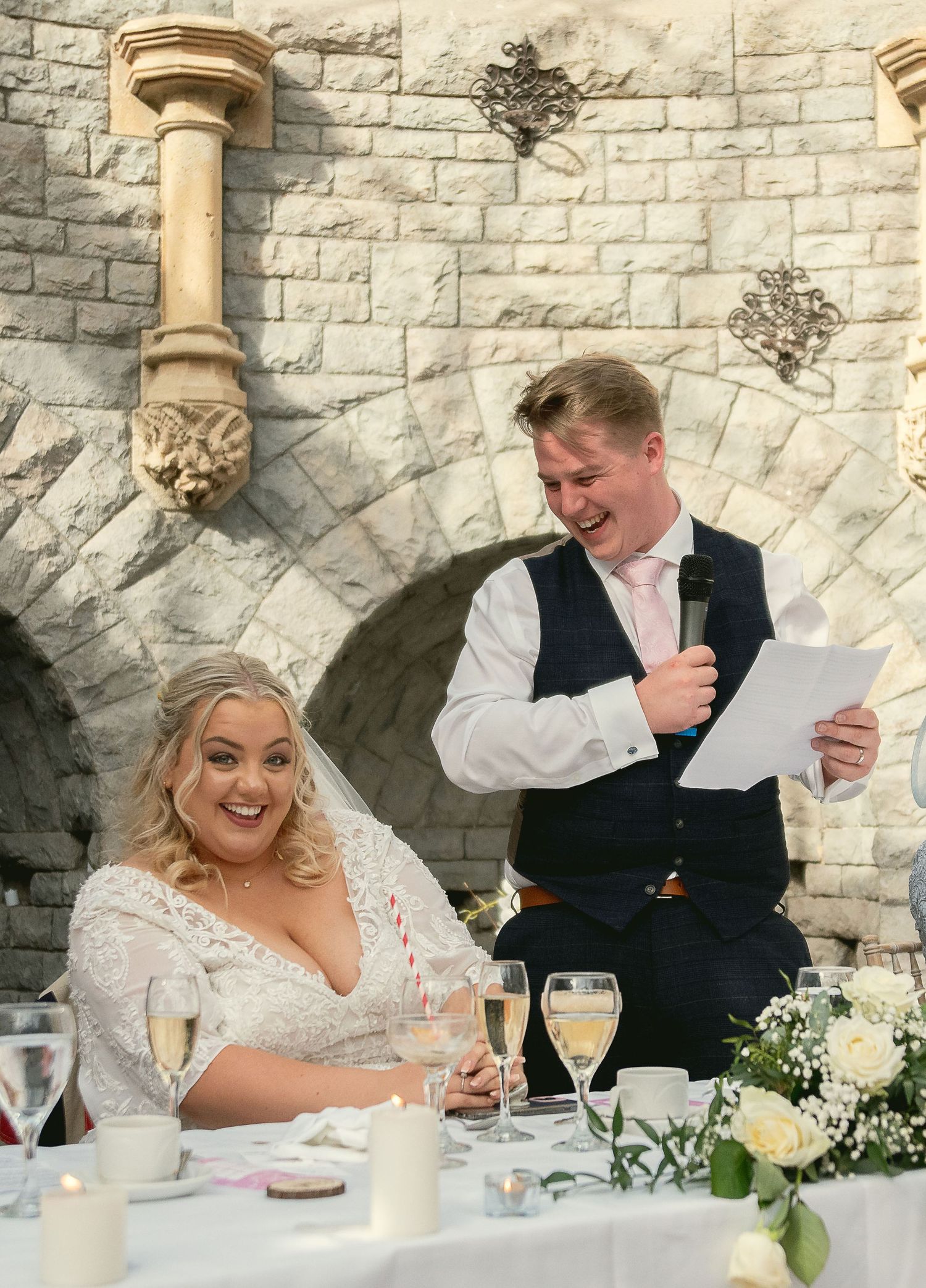 Wedding couple seated at head table reading speeches during reception in stone venue.