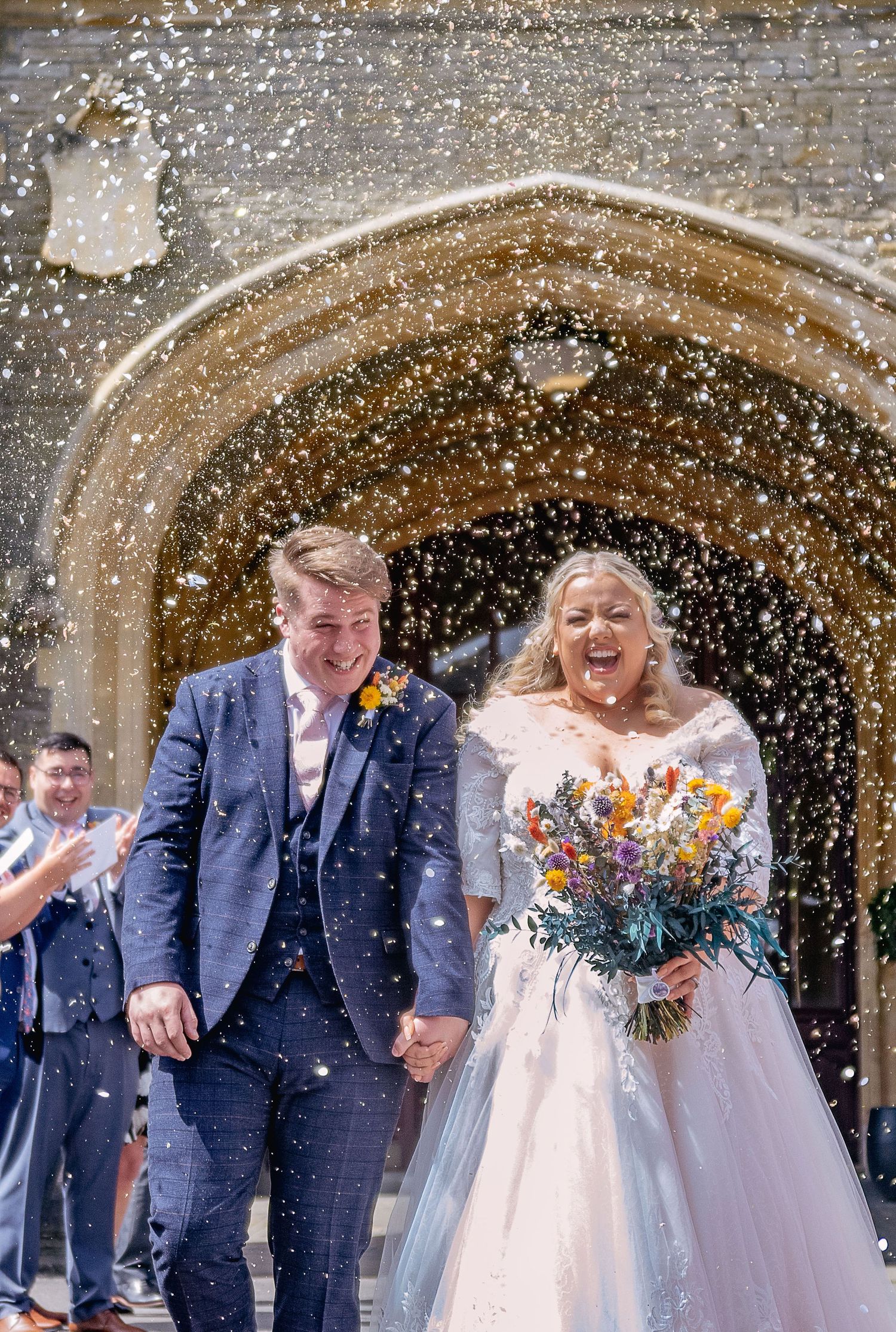 A joyful wedding moment captured as confetti falls around the couple under a stone archway.