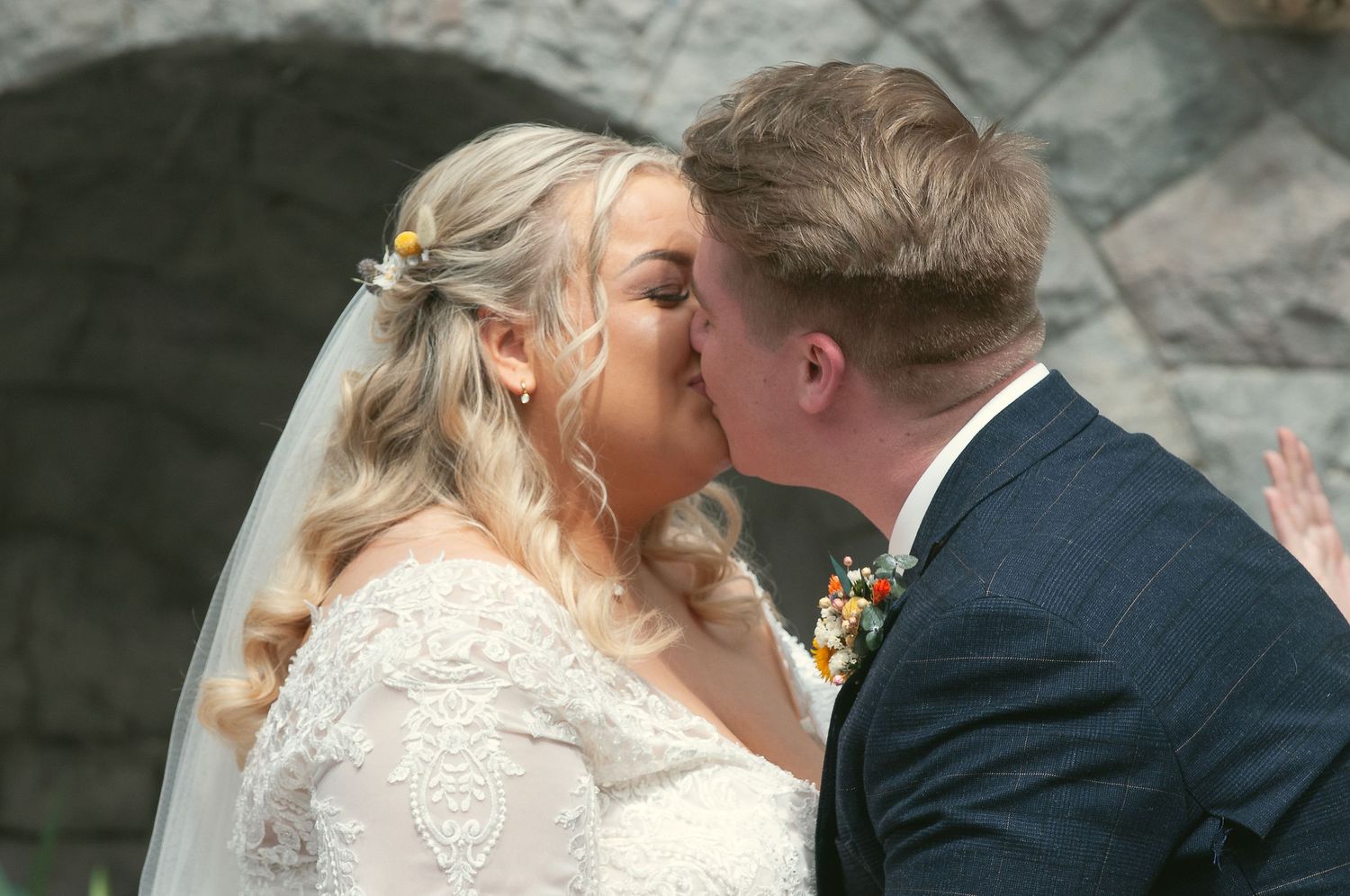 Close-up of newlyweds sharing a romantic kiss against stone architecture.