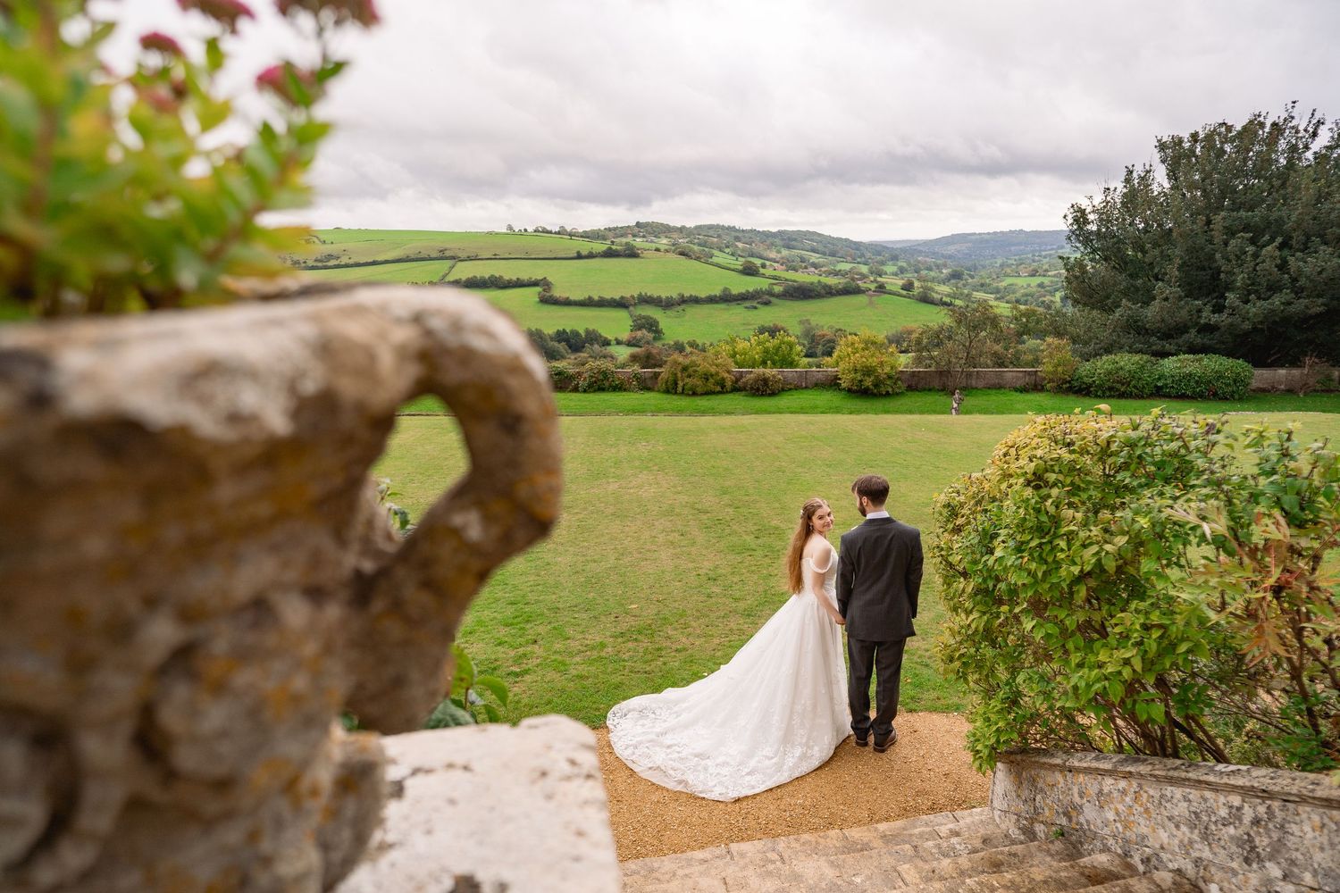 Bride and groom stand on a stone terrace overlooking lush green rolling hills at an elegant countryside wedding venue.