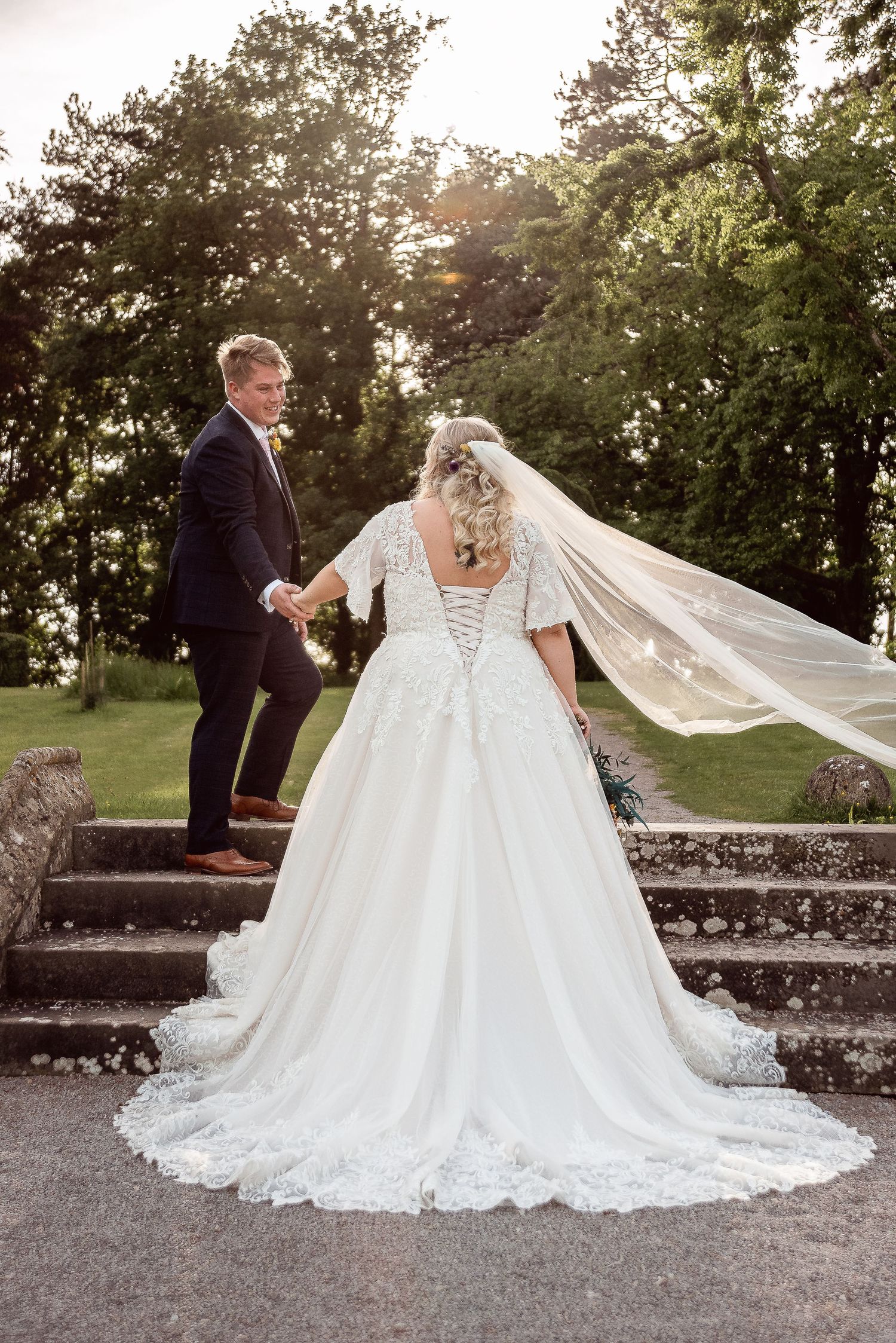 A flowing white wedding dress with lace details spreads beautifully across a gravel pathway at sunset.