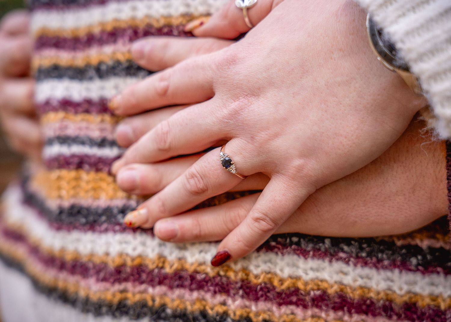 close up of hands joining during an embrace, focus on the emerald engagement ring. Jumper with warm toned stripes create colourful backdrop