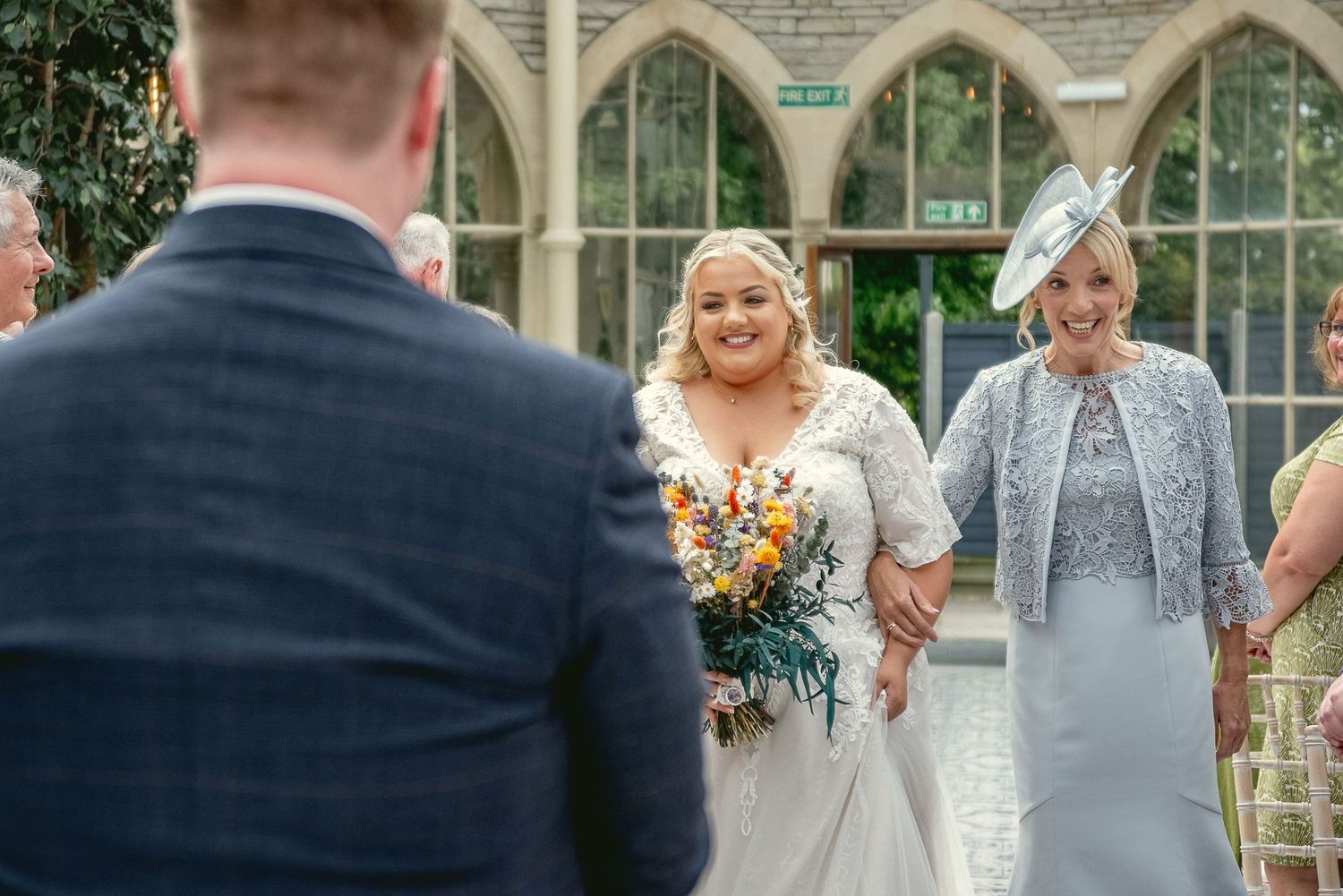 Bride in lace dress holding orange bouquet walking down aisle at gothic venue wedding ceremony.