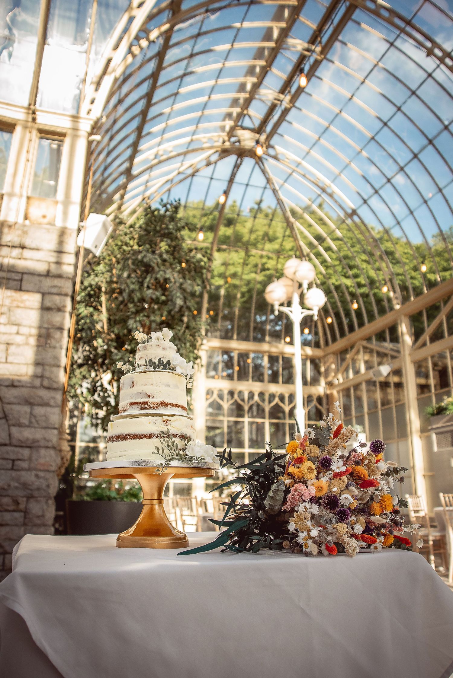 A conservatory wedding reception setup with glass ceiling and floral centerpieces on white tablecloths.