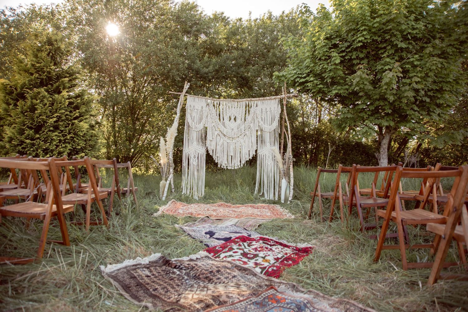Wide shot of rustic outdoor wedding ceremony setup with wooden chairs and macrame backdrop.