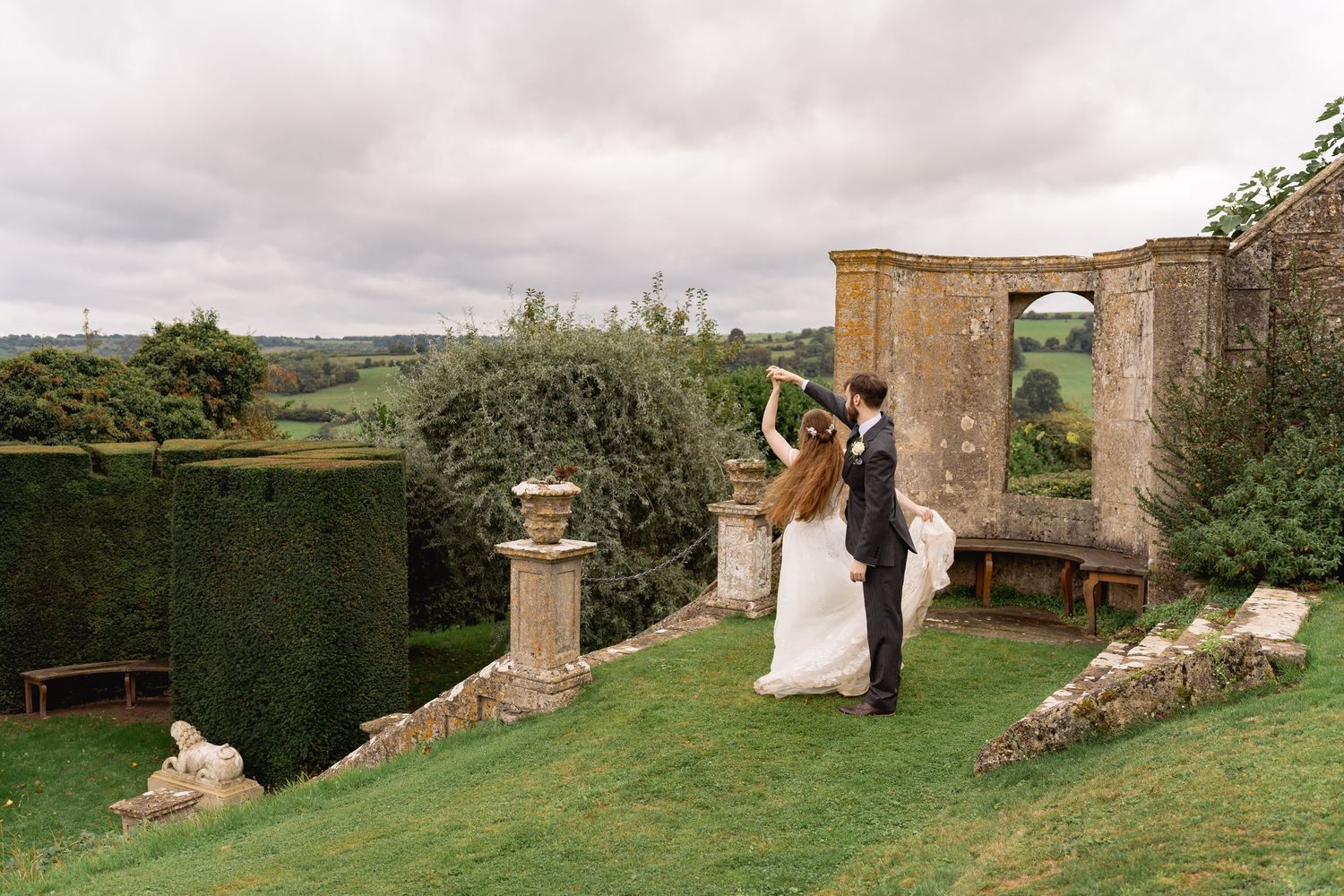 Bride and groom dancing on lush garden terrace with scenic countryside views and stone architectural ruins.
