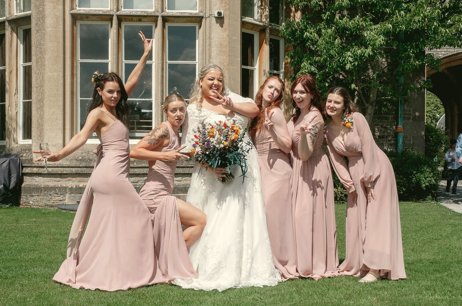 Bridal party in pink dresses striking fun poses on manicured lawn.
