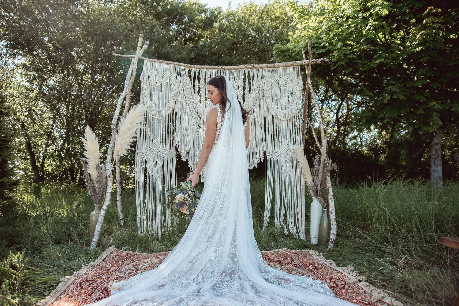 A bride in a lace wedding gown poses in front of a macrame backdrop in a woodland setting.