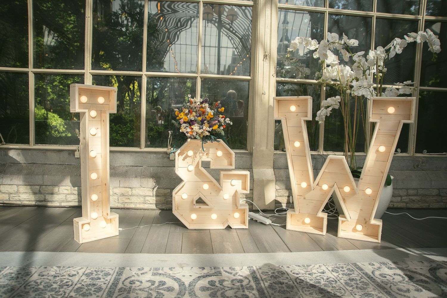 Illuminated wooden letters spelling L&W with floral decorations in a sunlit conservatory setting.
