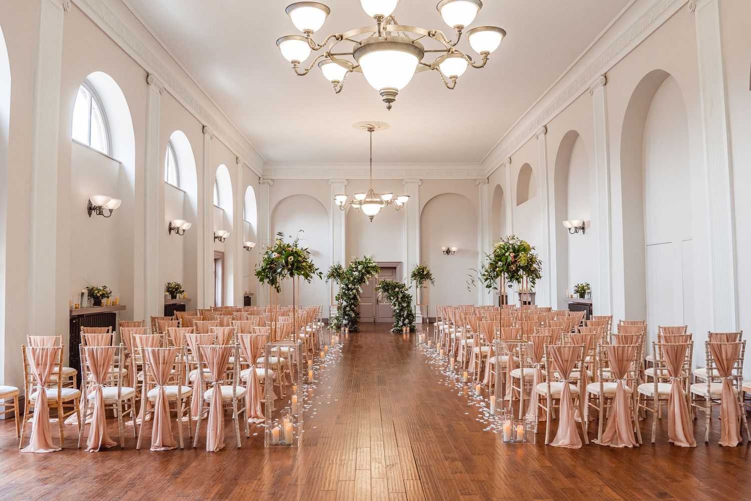 Ceremony set up in the Assembly Room. Bright room, tall ceilings and gold chandeliers. Rows of chairs with pink sashes.