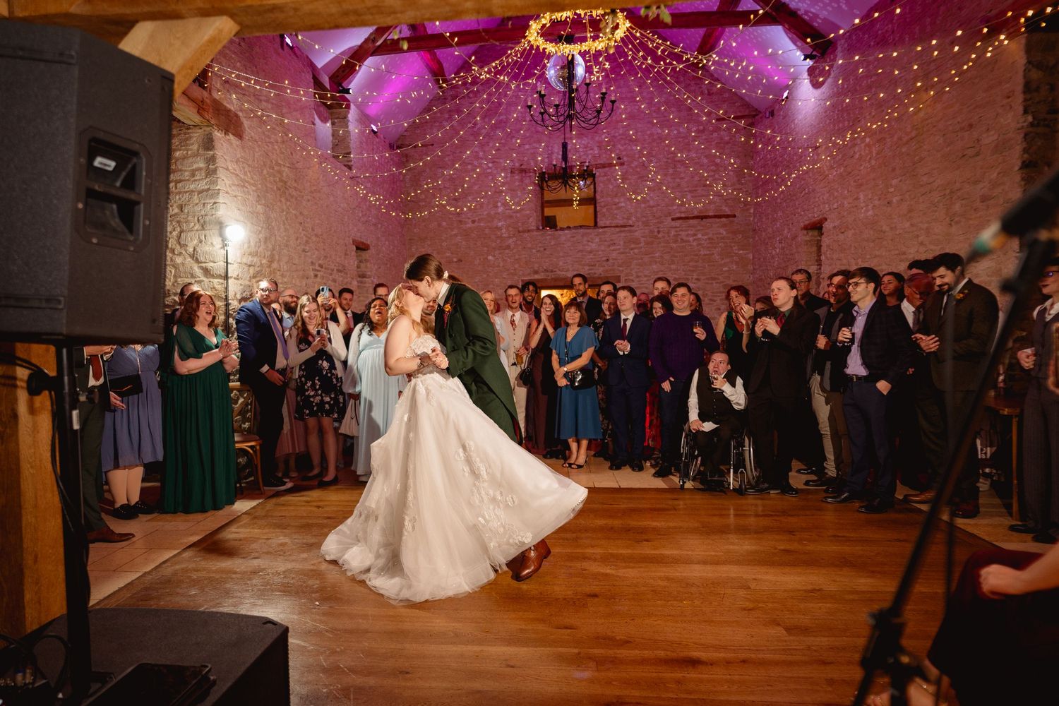 Bride and groom share first dance in rustic barn venue with fairy lights and purple uplighting, surrounded by wedding guests.