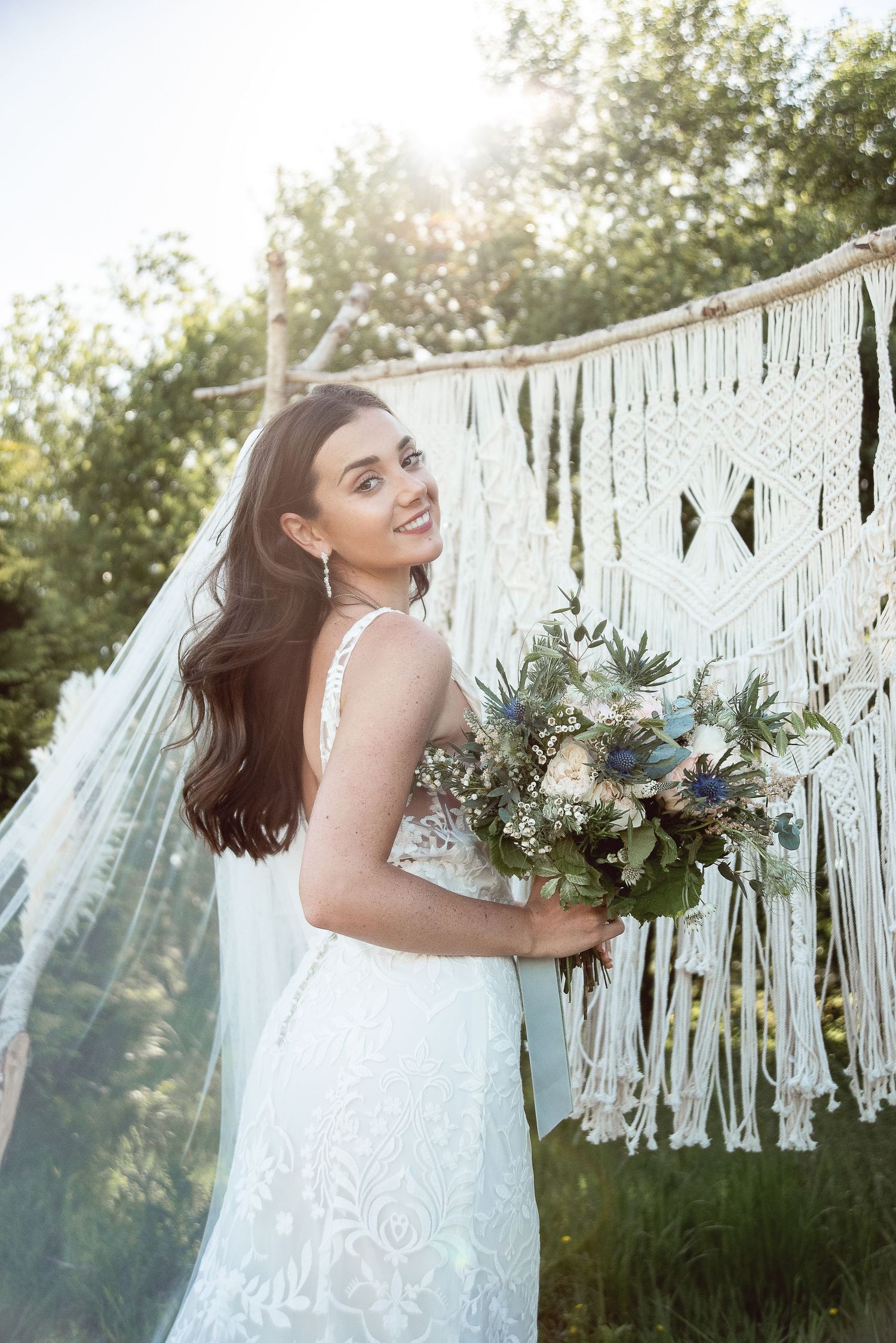 Sunlit bridal portrait featuring an elegant white dress and natural bouquet against a bohemian macrame backdrop.