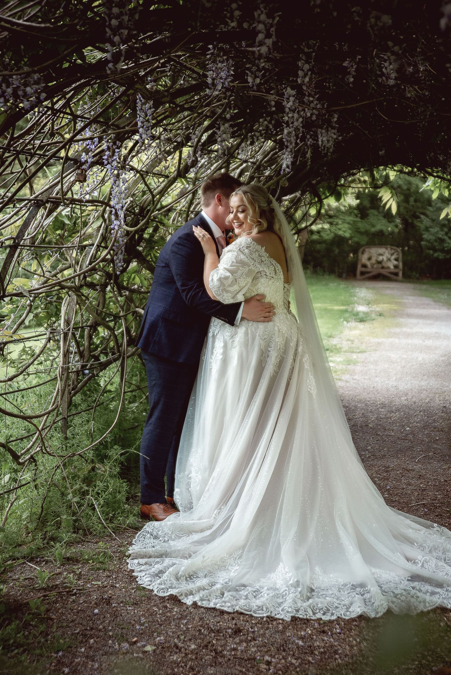 A romantic wedding photo captured under overhanging tree branches on a garden path.