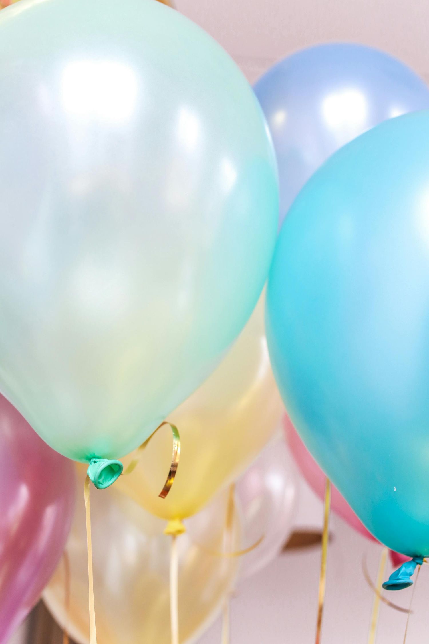 Colorful pastel party balloons in mint green, blue, and pink tones with gold ribbons against a light background.