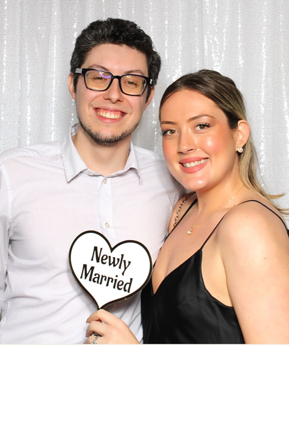 A happy couple poses in a photo booth with heart-shaped 'Newly Married' signs against a white backdrop.