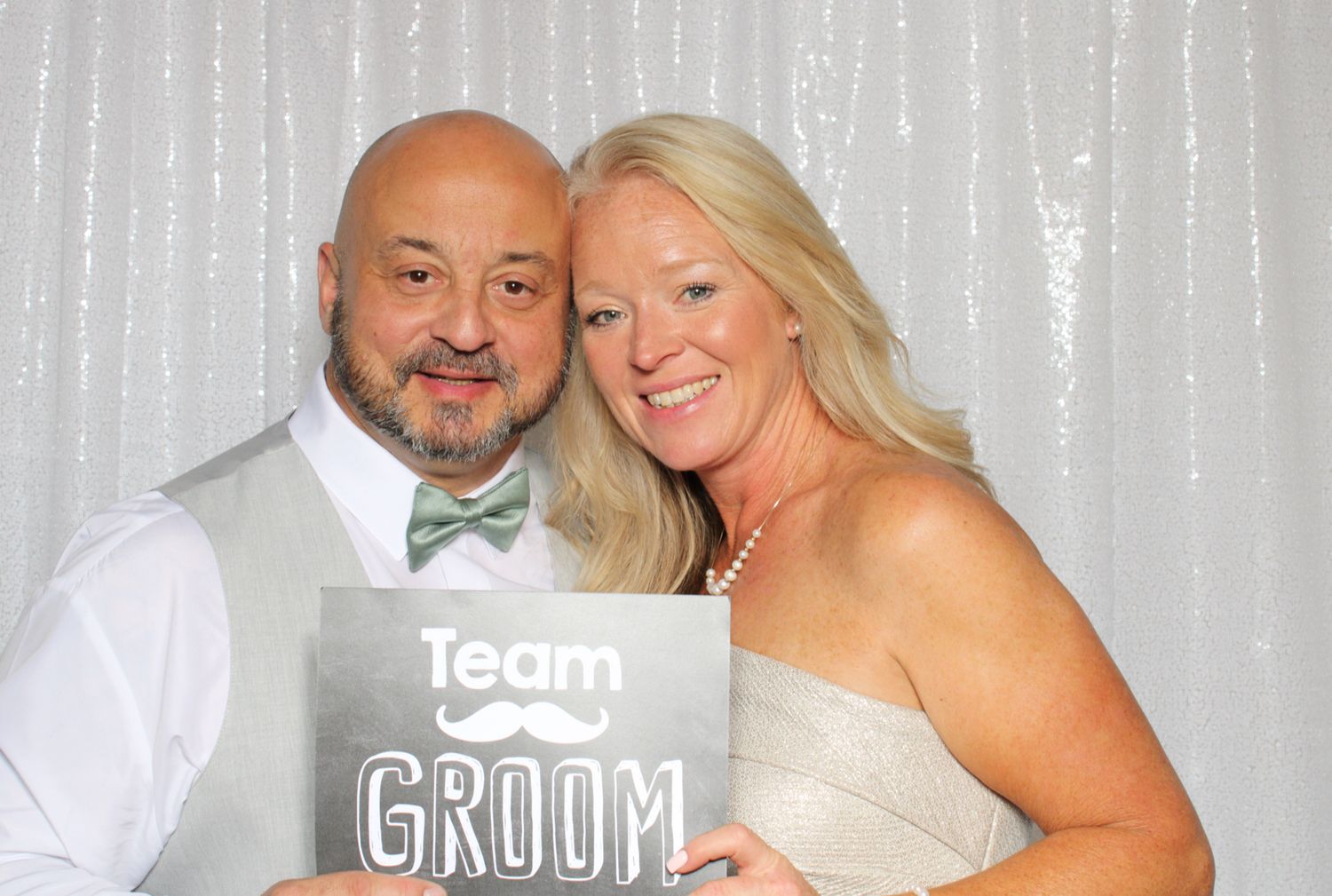 Couple poses with Team Groom sign in front of silver curtain backdrop.