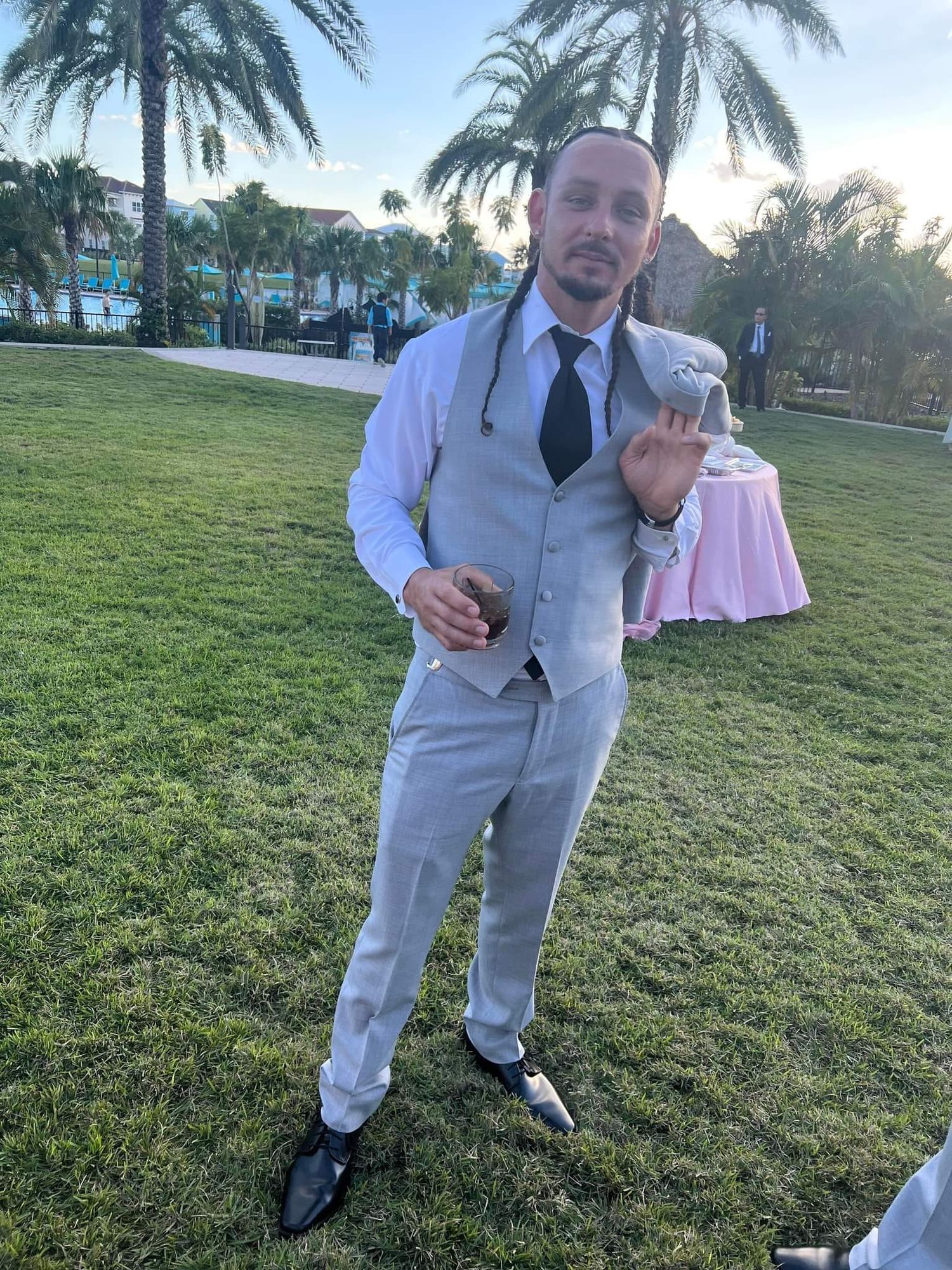 A person in a light gray suit poses playfully on a grassy lawn with palm trees in the background during sunset.