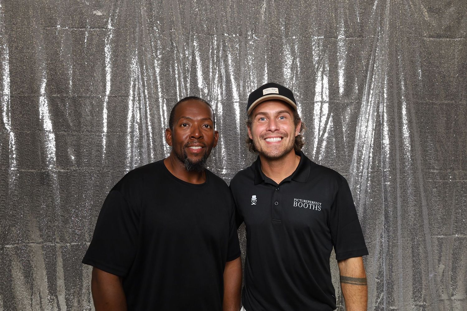 Two men in black shirts smiling together in front of a silver metallic backdrop at an event.