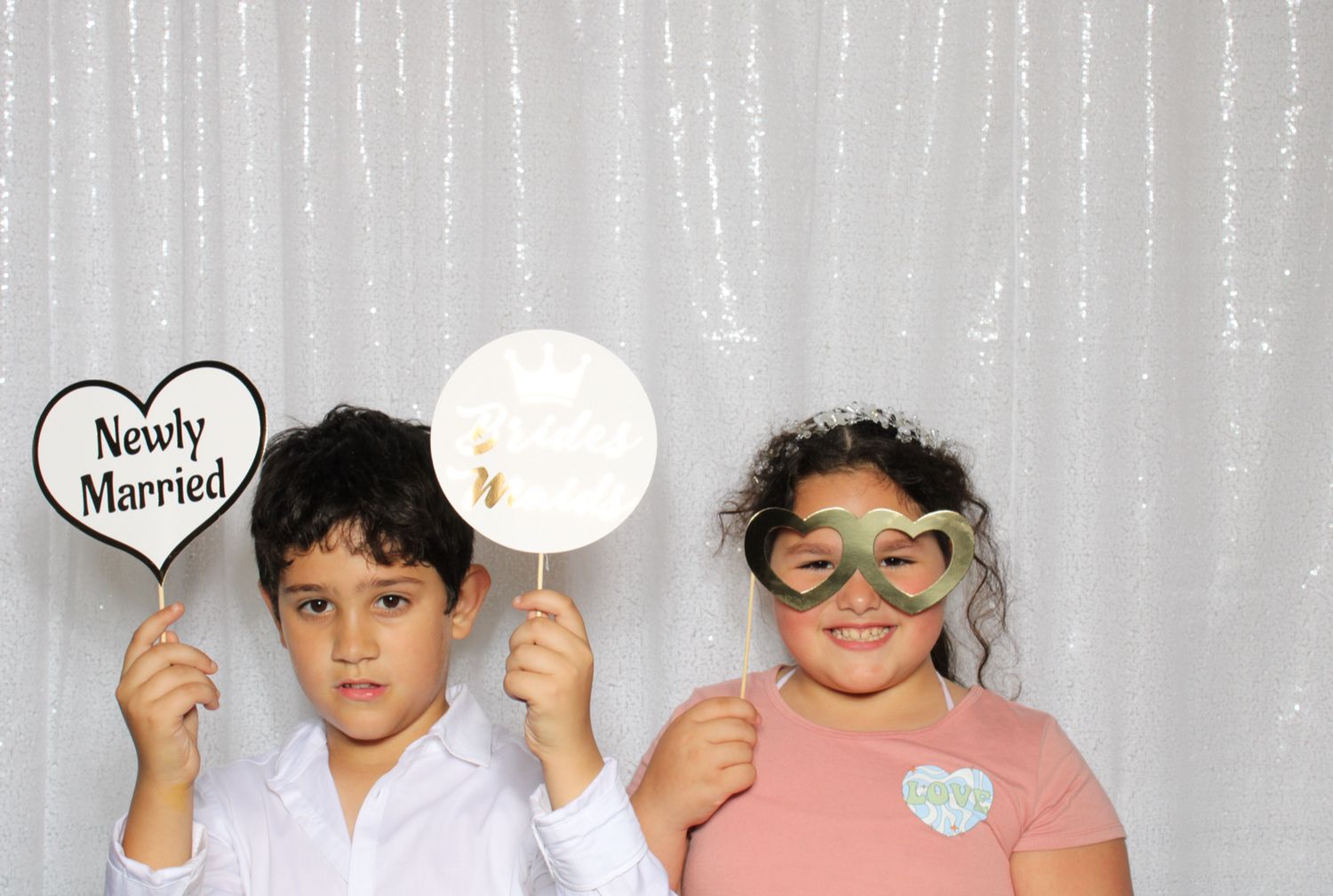 Two children pose at a wedding photo booth holding a Newly Married sign and decorative props with a white backdrop.