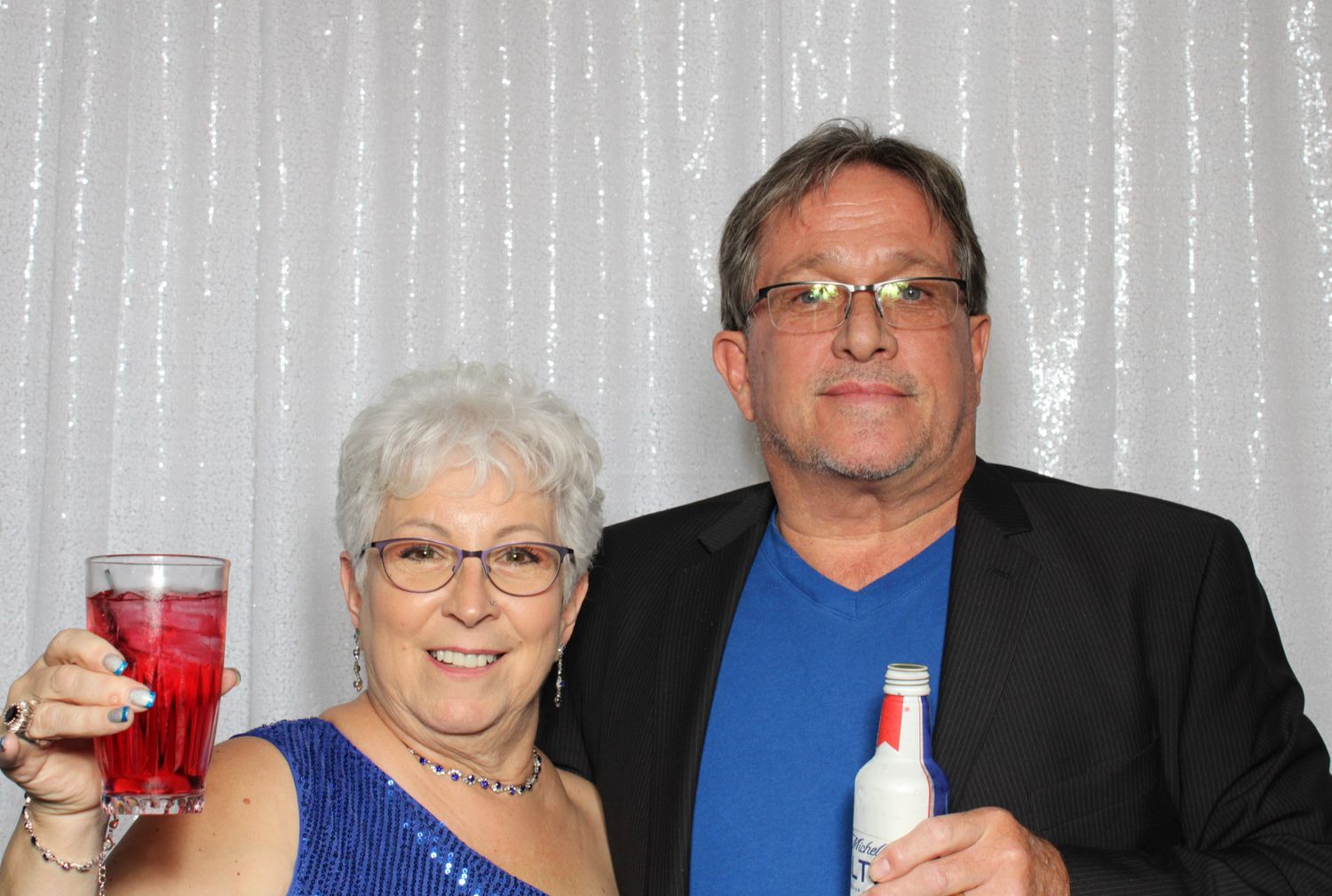 Couple at a party photo booth holding drinks and smiling in front of a silver curtain backdrop.