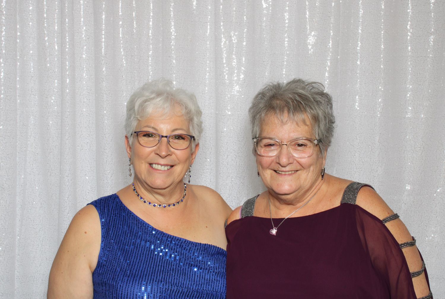 Two smiling older women in elegant blue and burgundy dresses pose together in front of a white sequin backdrop.