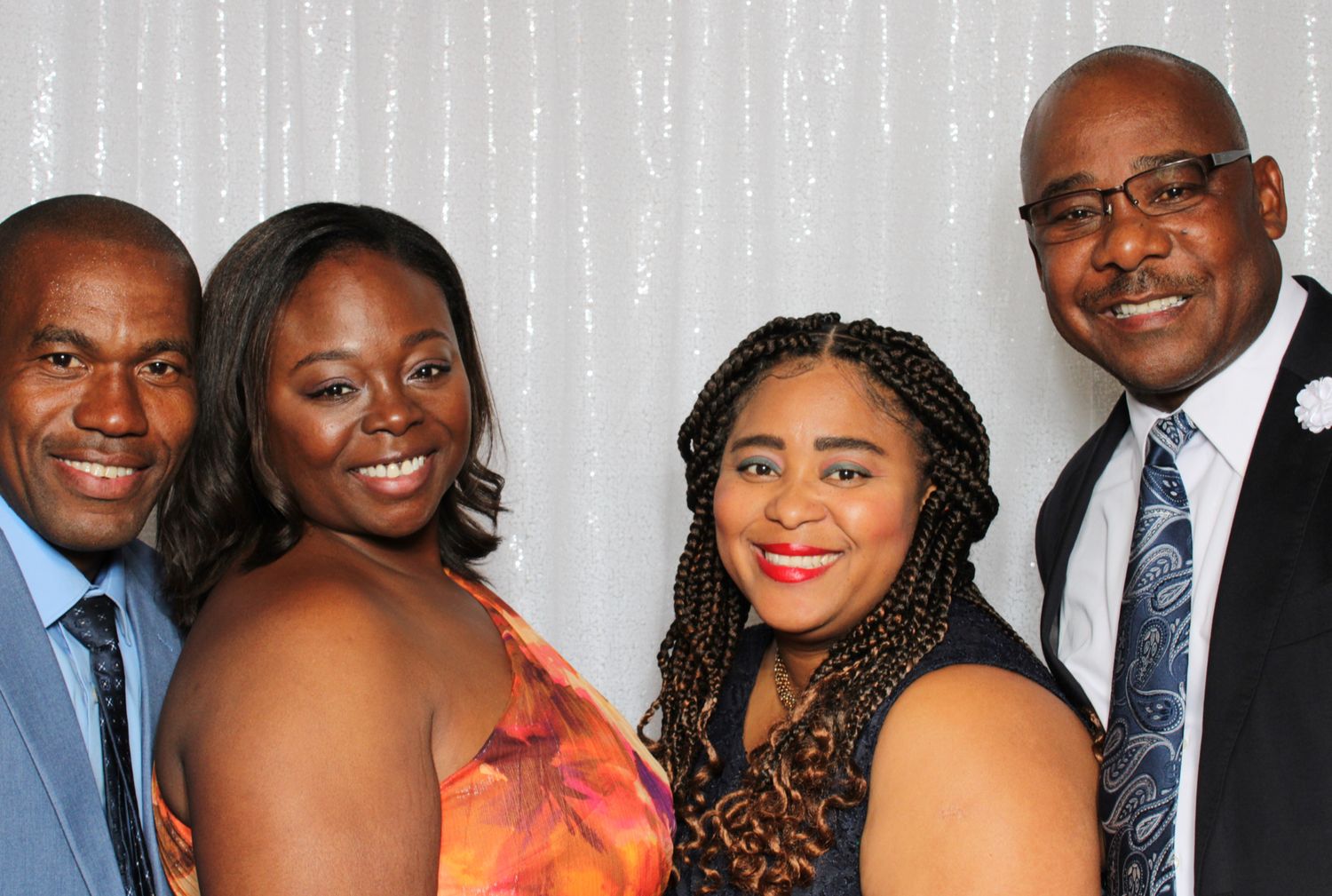 Two couples in formal attire pose together against a sparkly silver backdrop.