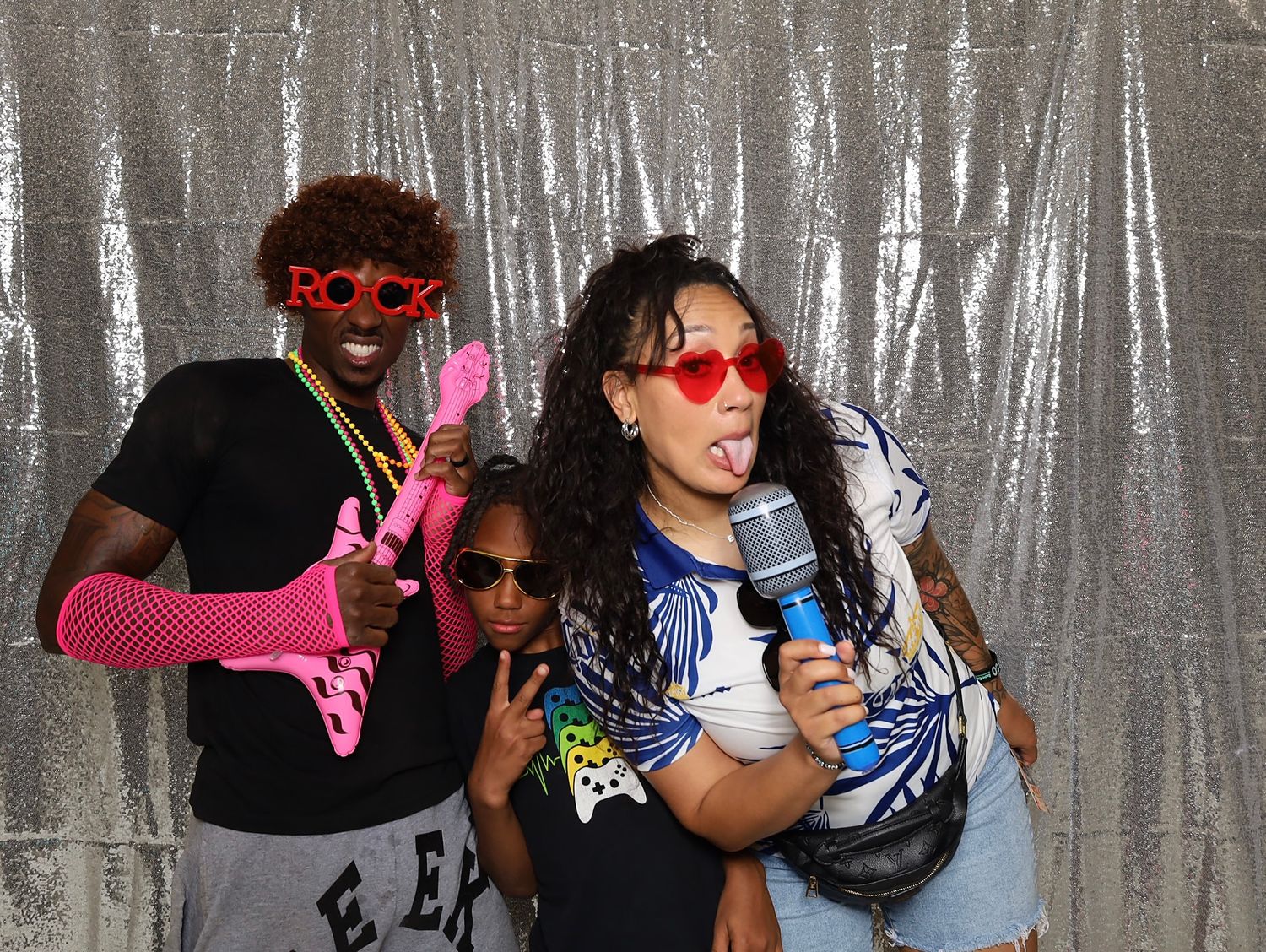 Three friends pose playfully in a photo booth with sequin backdrop, wearing fun accessories like heart sunglasses and pink props.