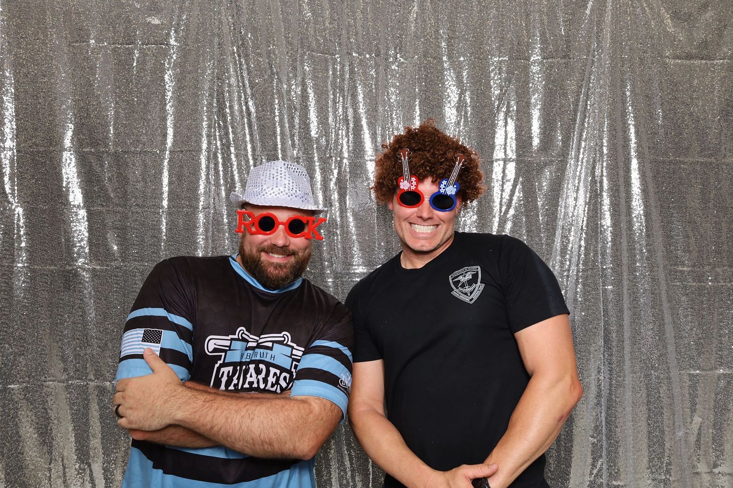 Two people wearing fun novelty sunglasses and hats pose together in front of a silver backdrop at a party event.