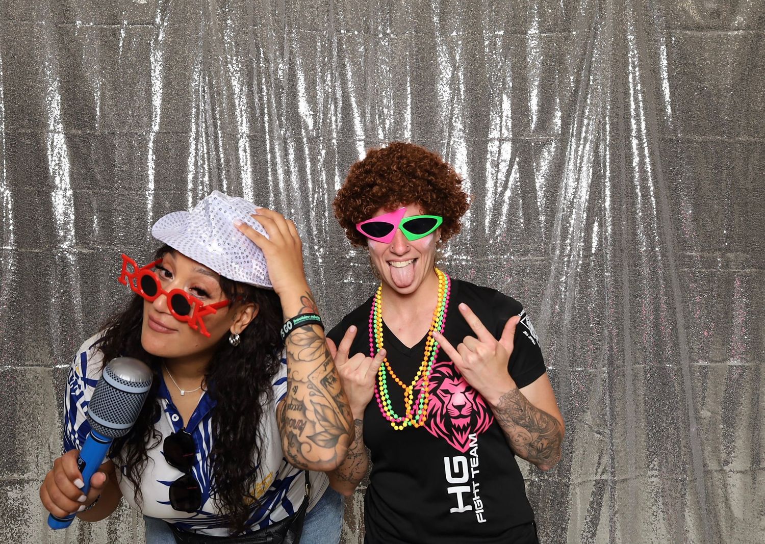 Two people in fun costumes with colorful glasses and accessories posing in a photo booth with silver backdrop.