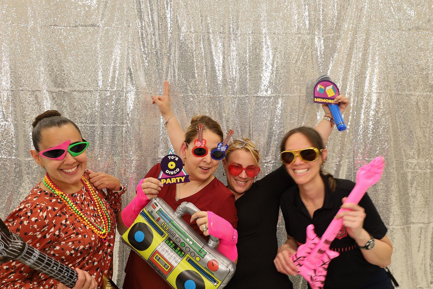 Four women posing in a photo booth with colorful props including a boombox, guitar, and fun sunglasses.
