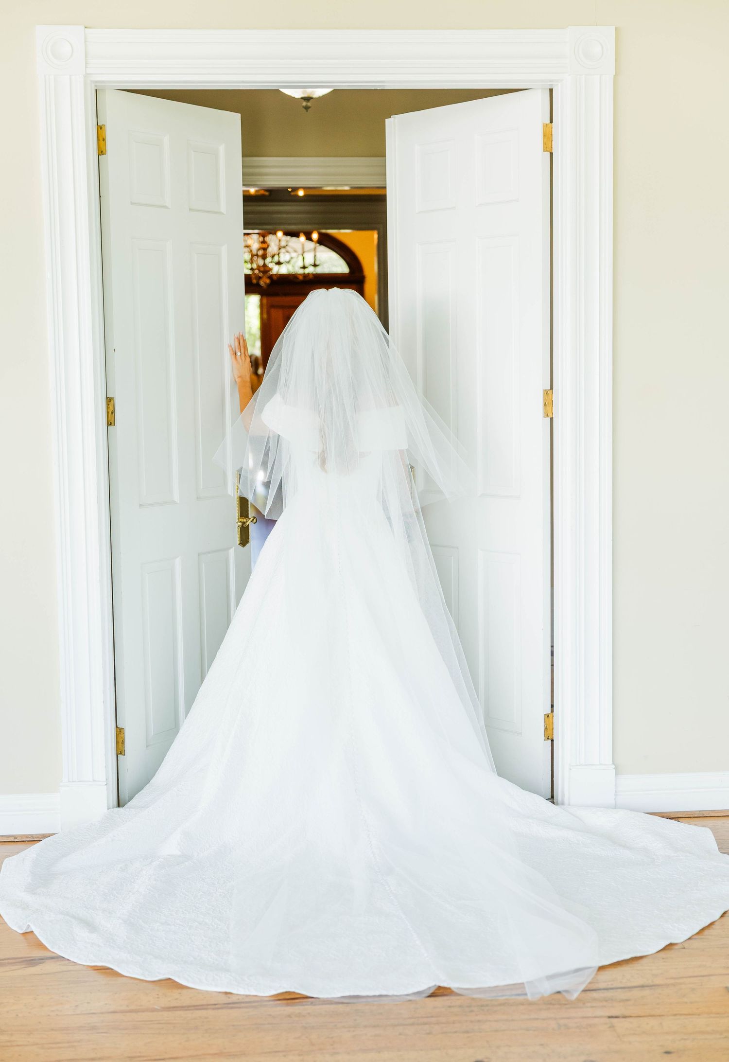Bride in white wedding gown with long train and veil standing in doorway, viewed from behind.