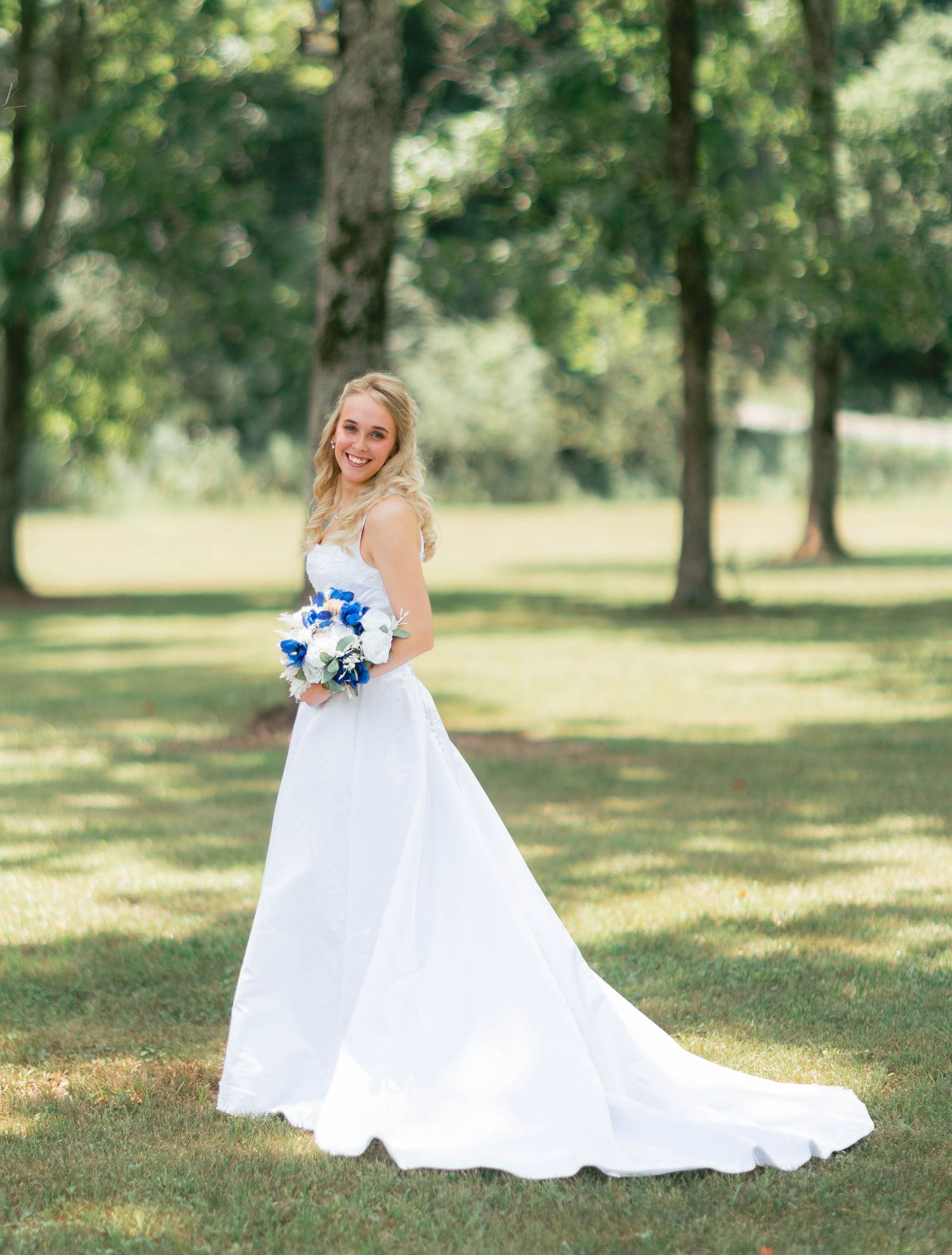 Bride in white gown holding blue and white bouquet poses outdoors in lush green park setting.