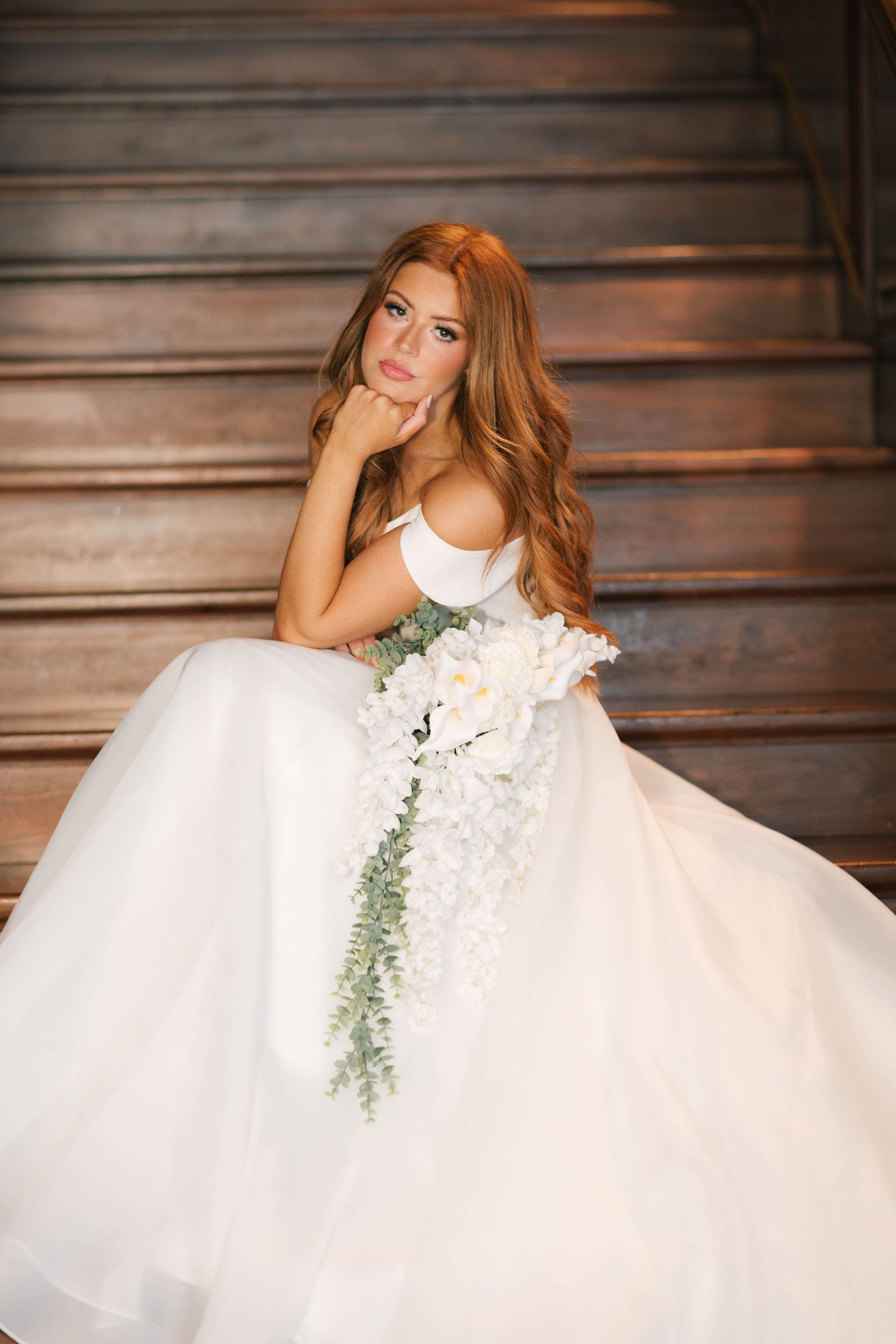 Bride in white ball gown holding cascading white floral bouquet, seated on wooden stairs with long red wavy hair.