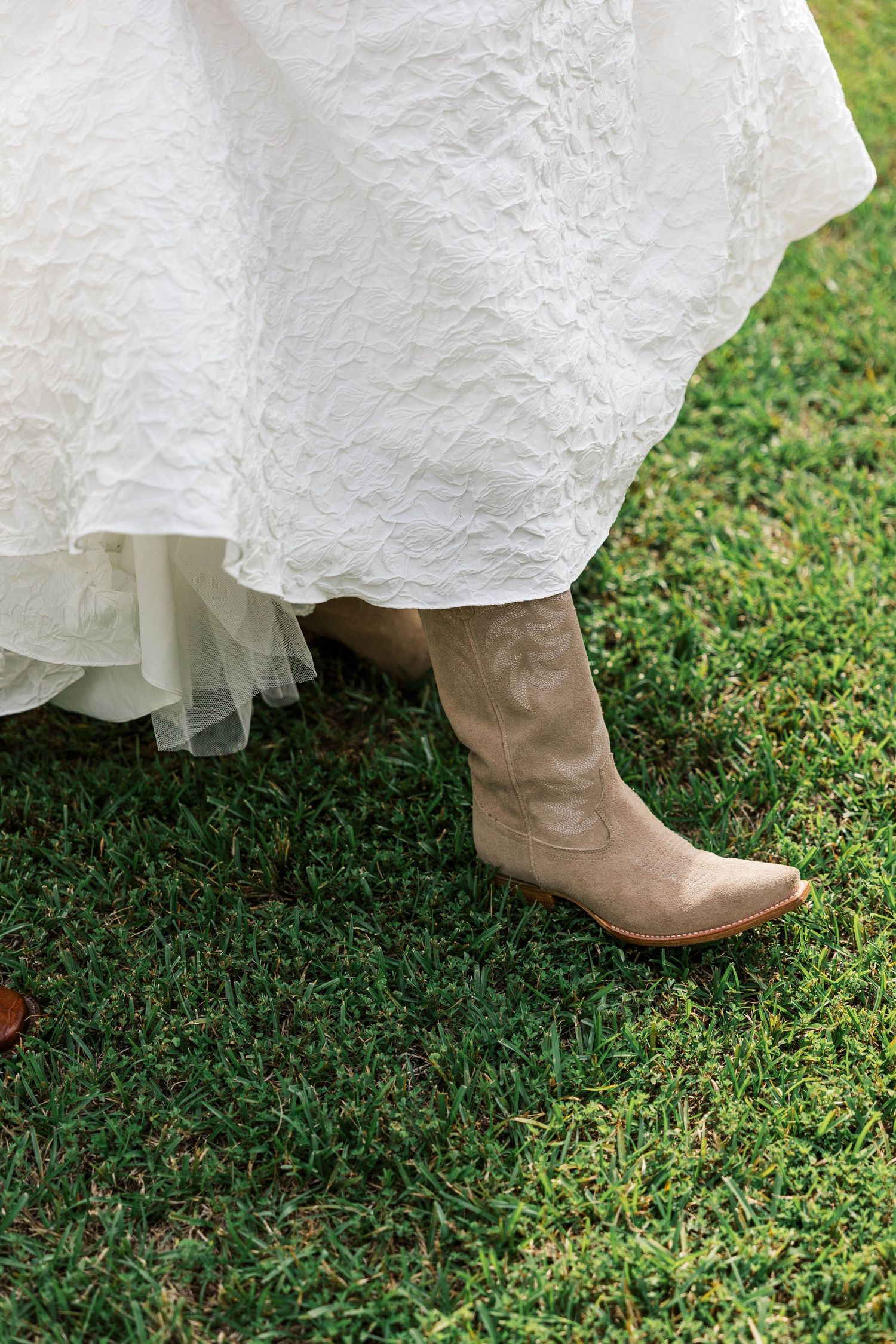Bride in white wedding dress and tan cowboy boots standing on green grass at outdoor wedding.