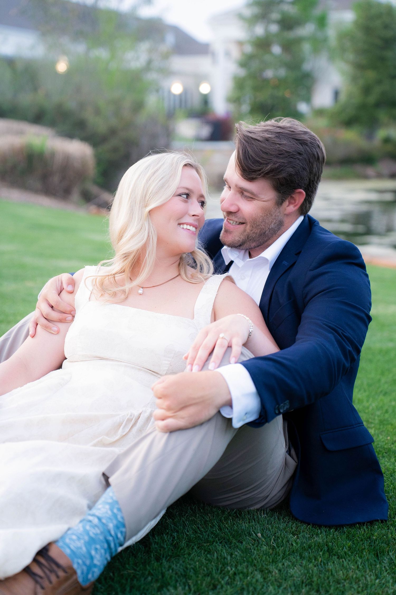 Couple sitting on grass smiling at each other, woman in white dress, man in navy suit, elegant venue background.