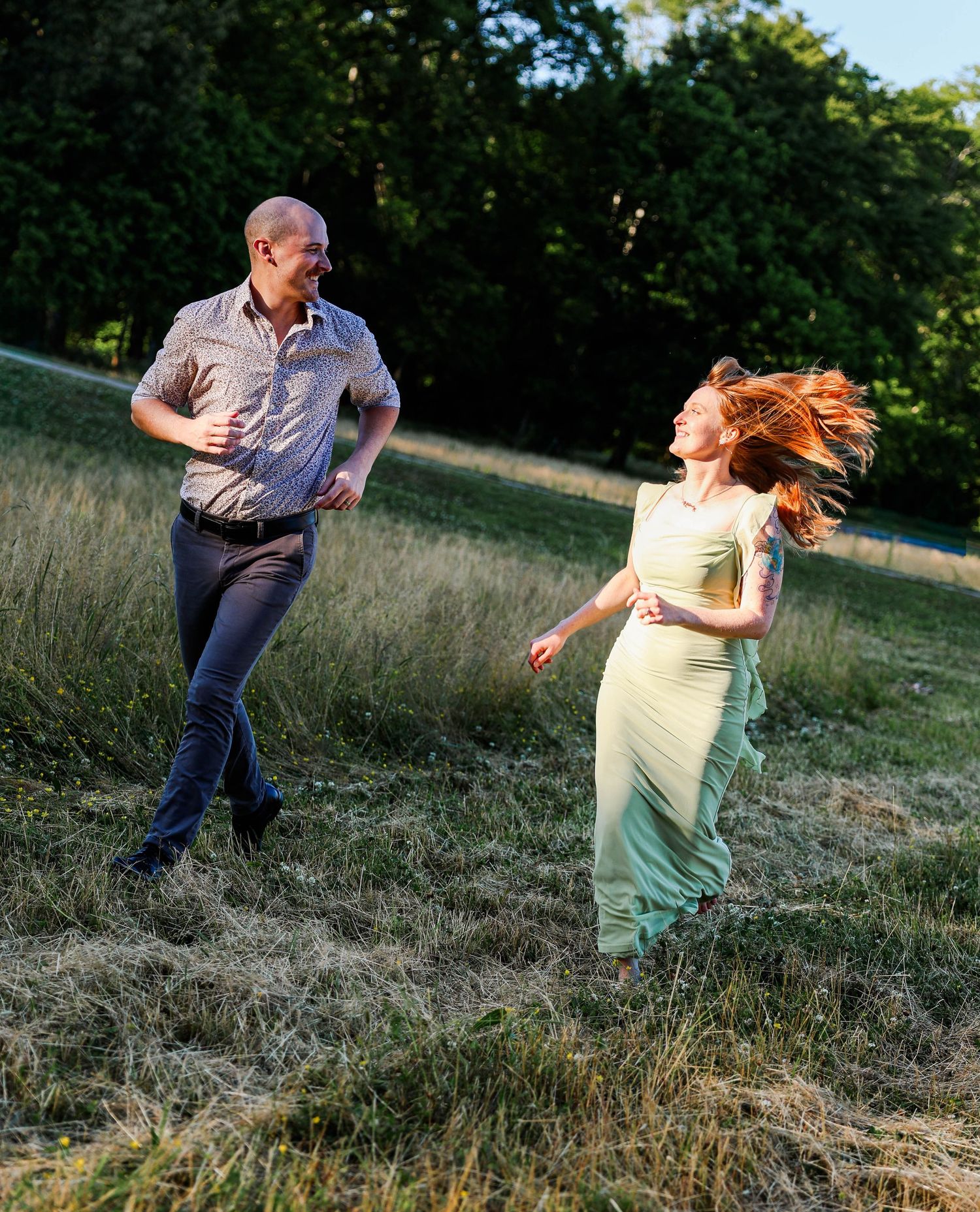 A man and woman run joyfully through a sunlit meadow, her red hair flowing, wearing a green dress.