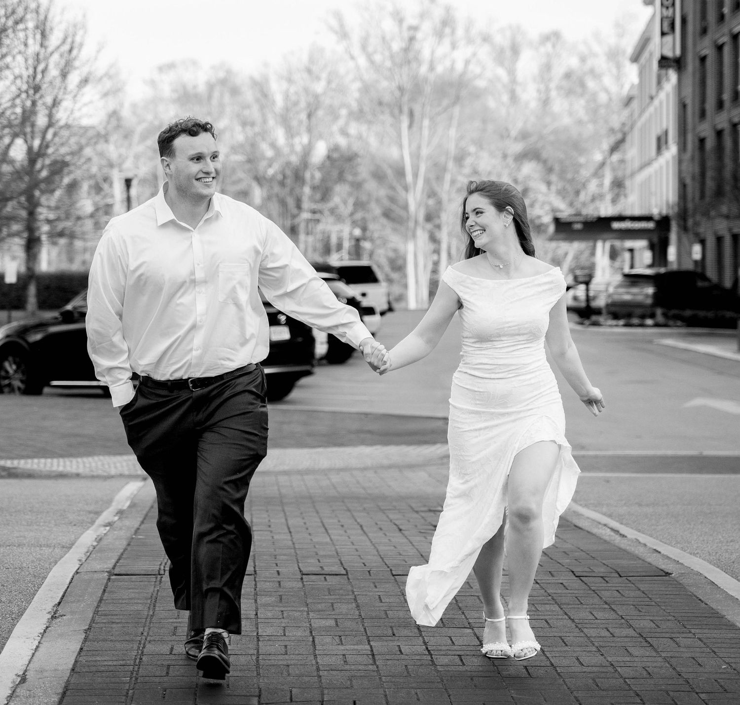 A joyful couple holds hands and walks on a brick street in black and white engagement photo.