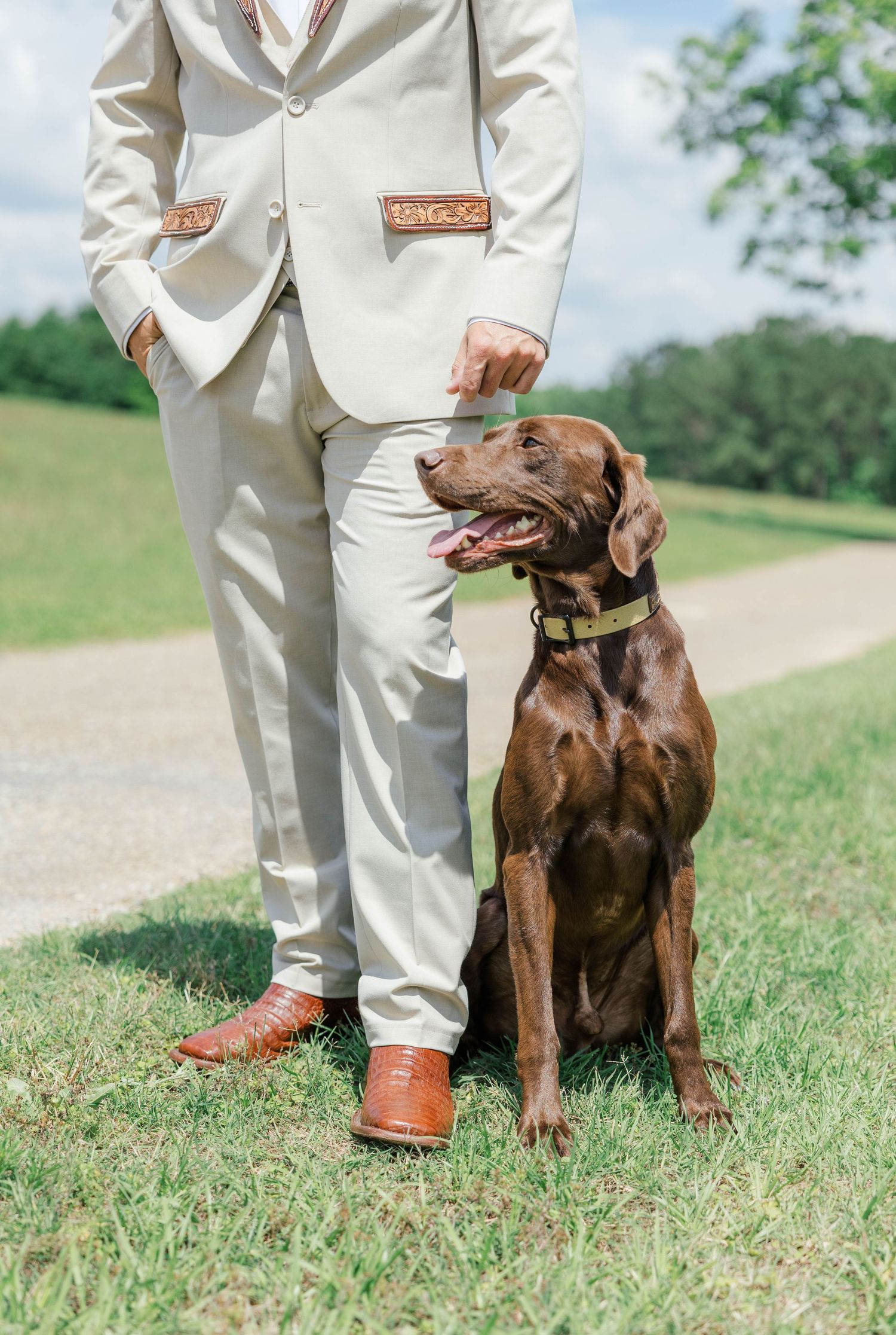 Groom in beige suit with brown cowboy boots stands beside a chocolate Labrador on green grass.