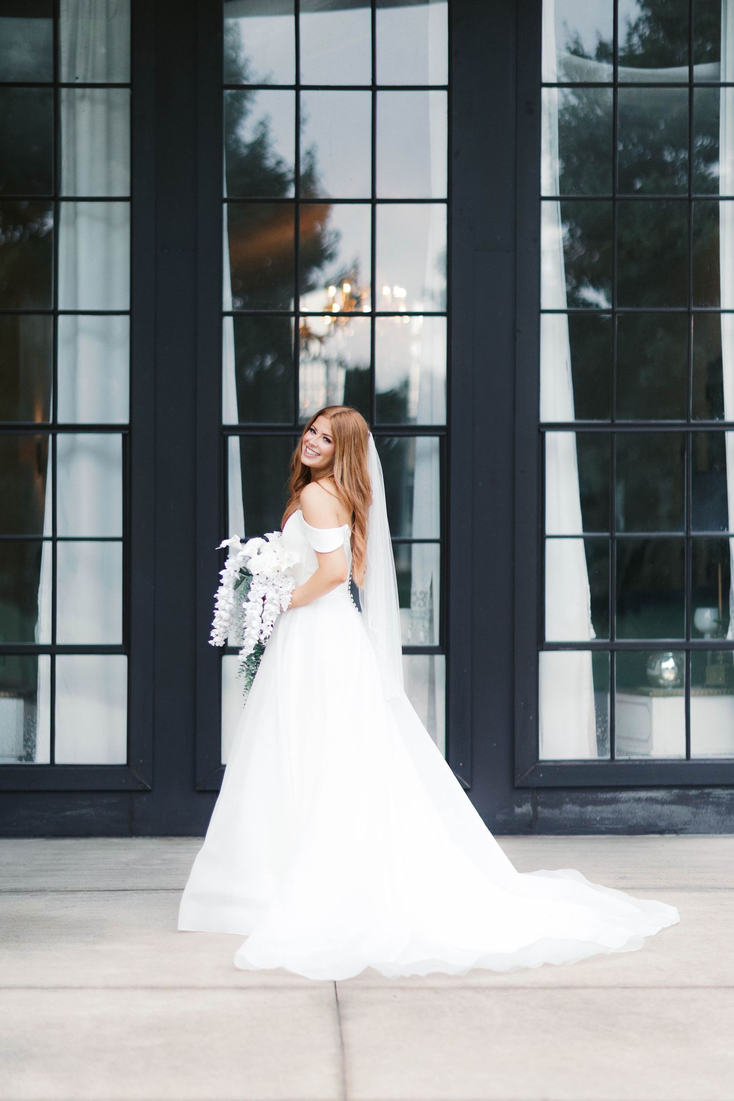 Bride in white off-shoulder gown with veil holds bouquet, posing before dark French doors.