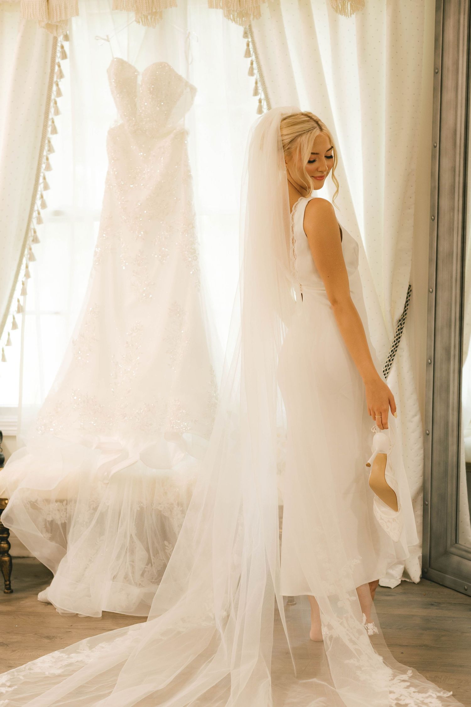 Bride in fitted white gown with long veil stands near draped window beside hanging lace wedding dress.