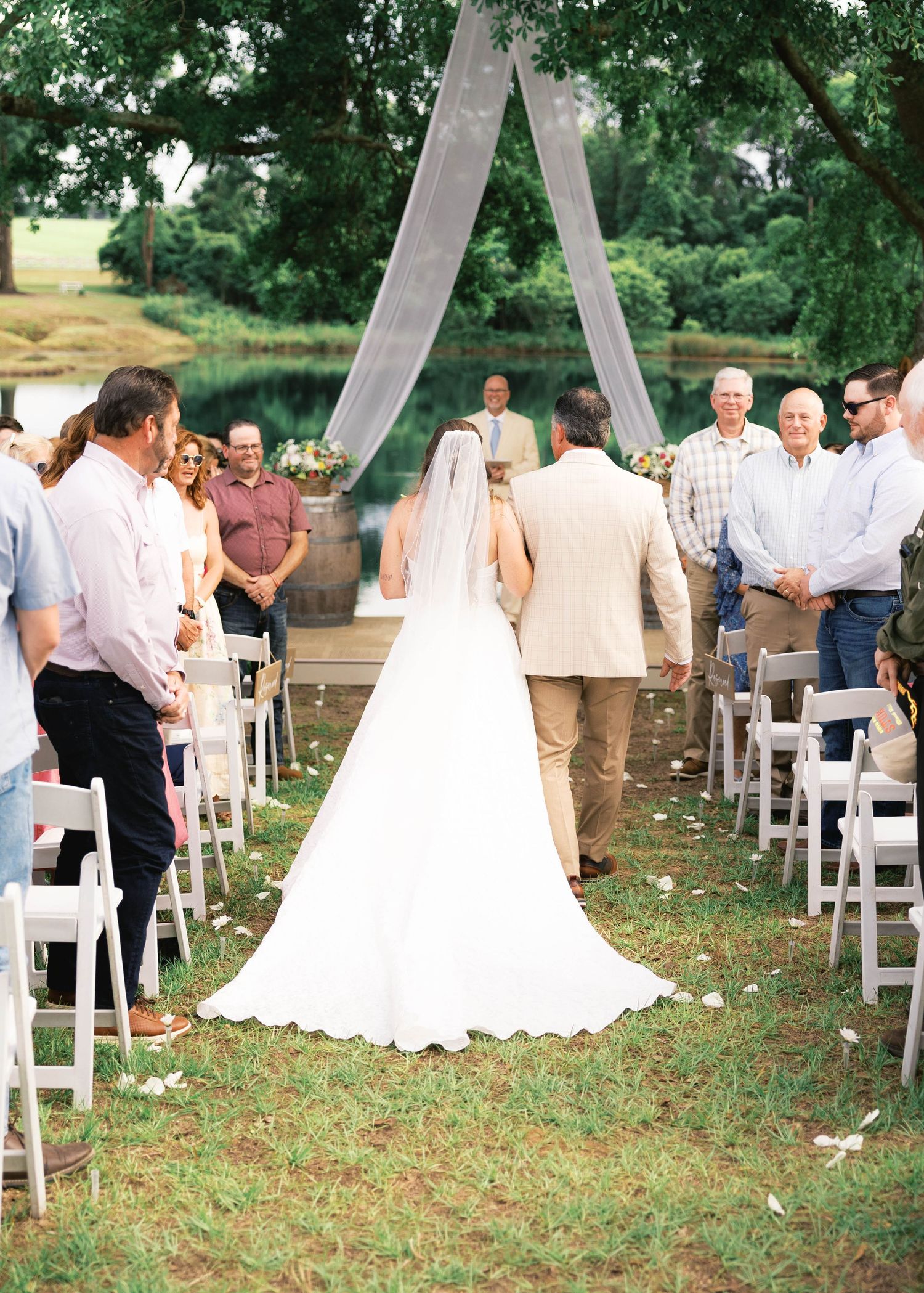 Bride walking down outdoor aisle with long veil, guests standing beside white chairs, sheer arch and pond backdrop.