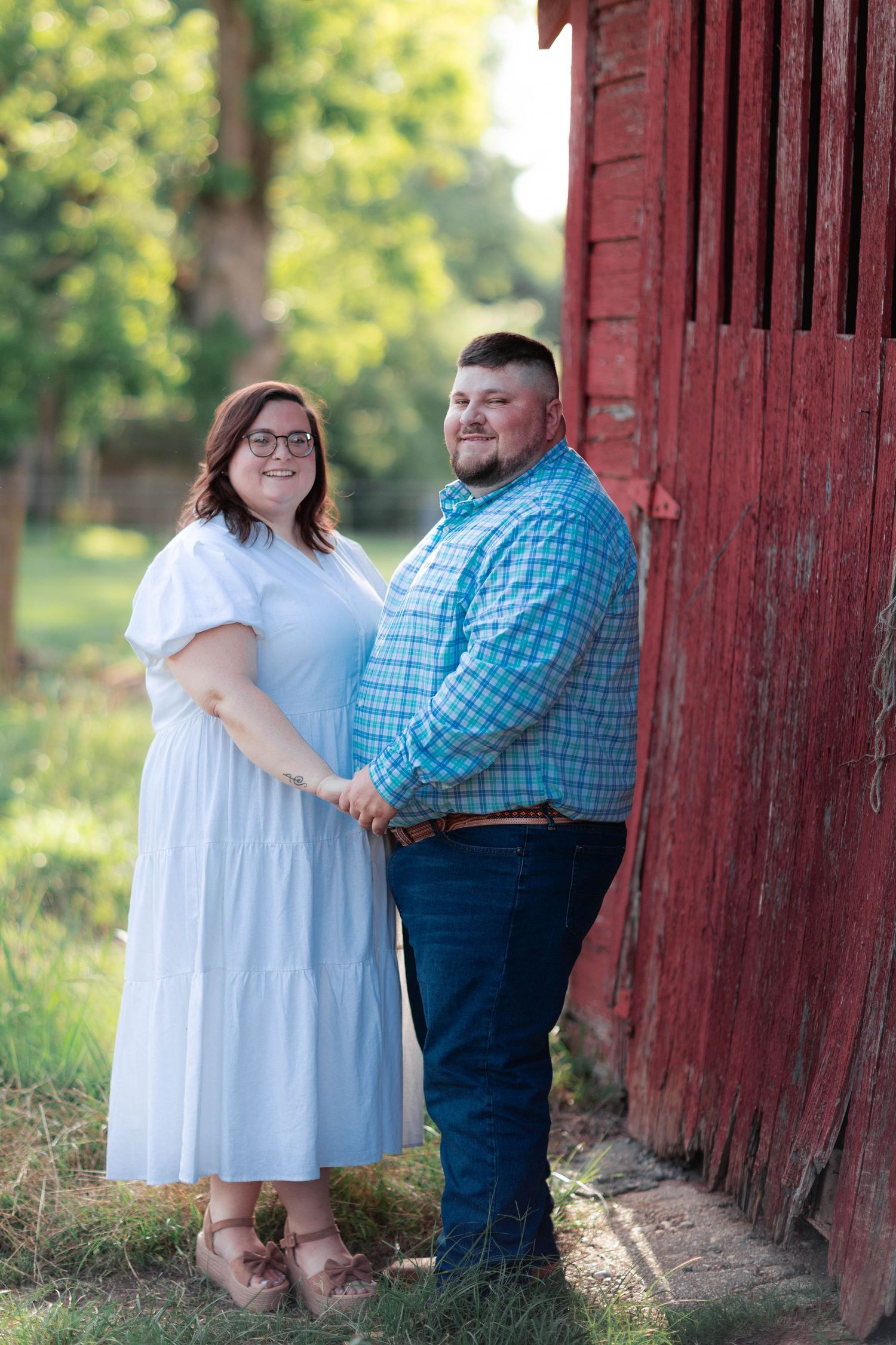 Couple holding hands smiling near a rustic red barn, woman in white dress and man in blue plaid shirt, outdoor portrait.