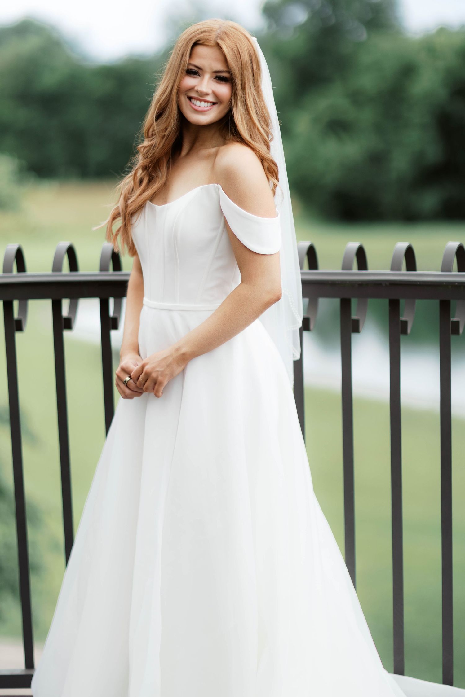 Smiling bride in off-shoulder white gown and veil stands on outdoor balcony with green trees in background.