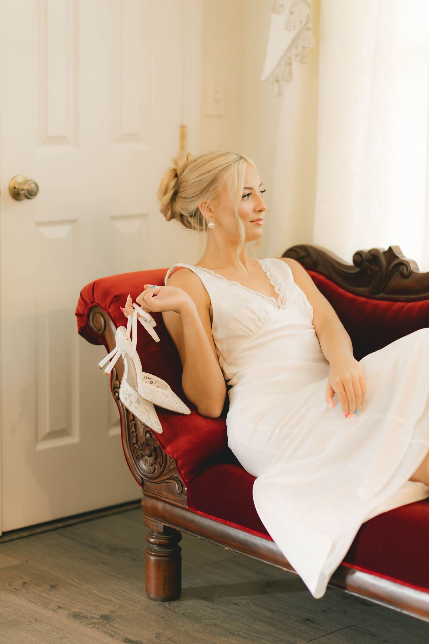 Smiling blonde bride in white wedding gown sits on red velvet sofa holding bridal heels.